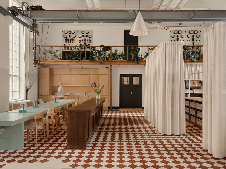 An expansive open-plan studio features a long wooden counter and a light teal work table set against a red and white checkerboard floor. An elevated mezzanine, adorned with lush plants, overlooks the space, blending industrial architecture with biophilic elements.