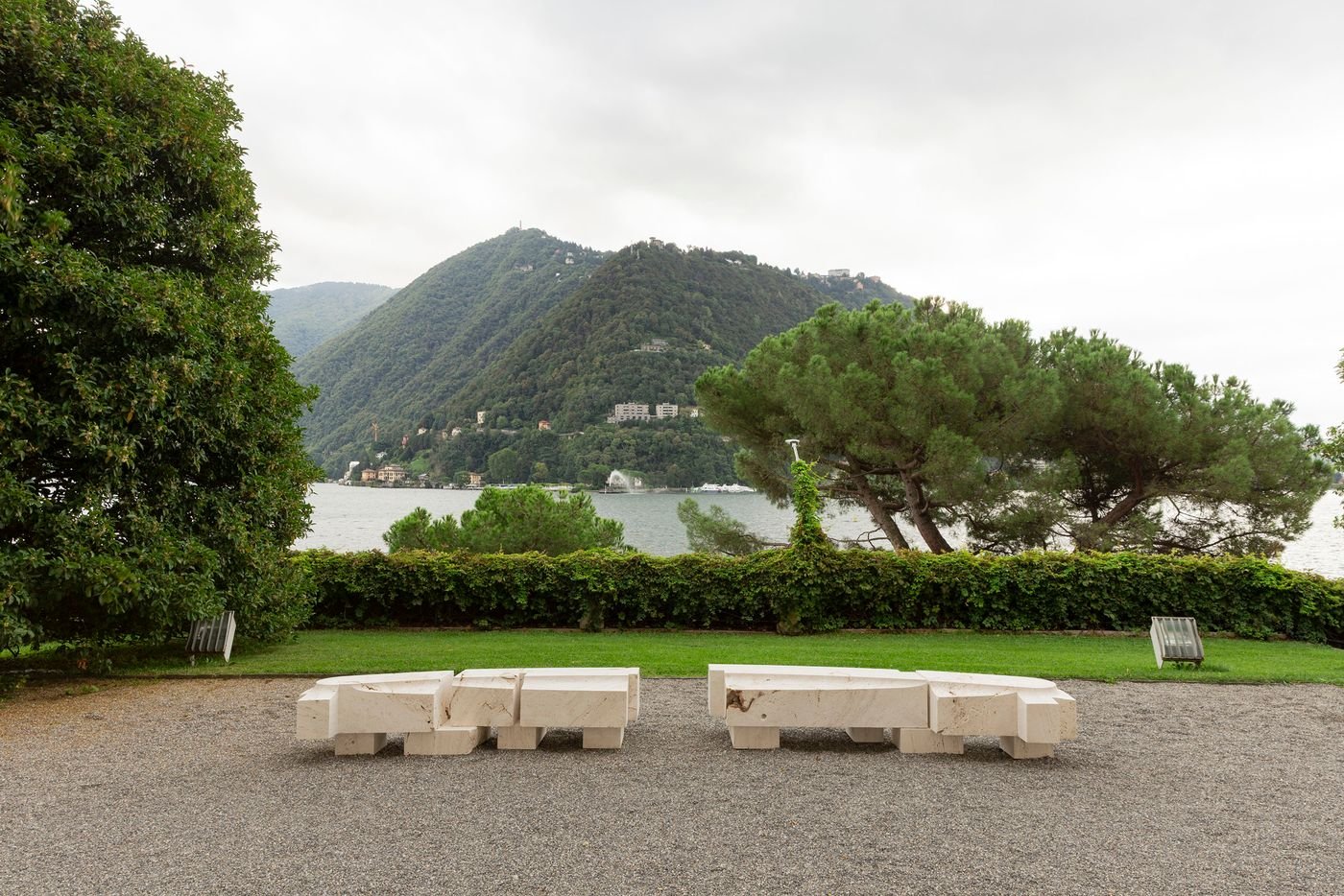 A pair of sculptural, light-colored stone benches are positioned on a gravel path, overlooking a serene Lake Como landscape. The benches are composed of stacked geometric blocks, their rough, unpolished texture contrasting with the smooth, manicured lawn. The composition frames a view of the still lake and the lush, tree-covered mountains, merging contemporary sculpture with the stunning natural environment.