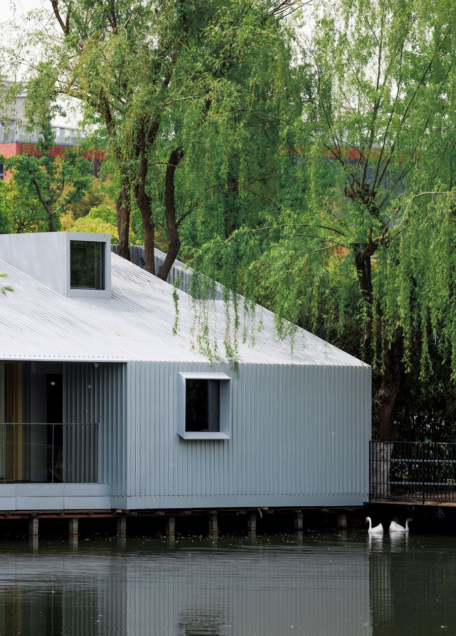 Wutopia Lab's Lake House extends over the water, its corrugated metal facade and gabled roof featuring a striking dormer window and a smaller square window. The structure’s clean lines contrast with the soft, flowing branches of weeping willows overhead and the gentle ripples of the lake below, where two white swans gracefully glide, highlighting the tranquil integration of modern architecture with the natural environment. Wutopia Lab's Lake House extends over the water, its corrugated metal facade and gabled roof featuring a striking dormer window and a smaller square window. The structure’s clean lines contrast with the soft, flowing branches of weeping willows overhead and the gentle ripples of the lake below, where two white swans gracefully glide, highlighting the tranquil integration of modern architecture with the natural environment.