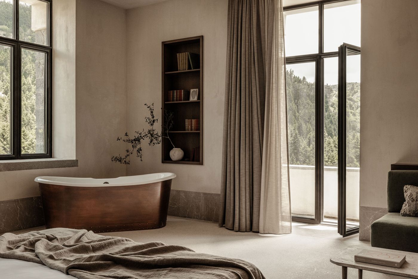 A luxurious and serene open-plan suite features a striking freestanding copper bathtub, positioned for a view of the natural landscape through a large window. Sheer and opaque drapes frame an expansive glass door leading to a balcony. A minimalist built-in bookshelf and a cozy green sofa complete this elegant, biophilic retreat. A luxurious and serene open-plan suite features a striking freestanding copper bathtub, positioned for a view of the natural landscape through a large window. Sheer and opaque drapes frame an expansive glass door leading to a balcony. A minimalist built-in bookshelf and a cozy green sofa complete this elegant, biophilic retreat.