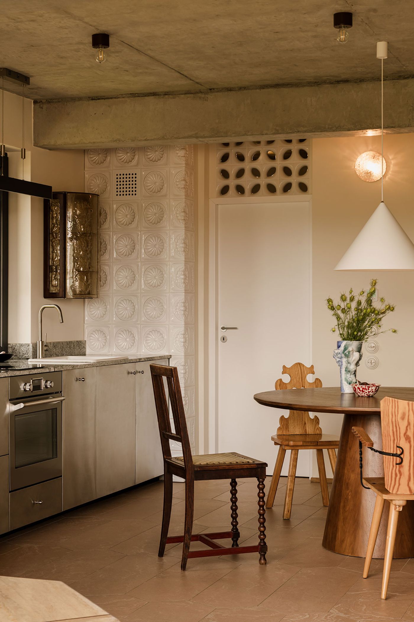 A modern kitchen with stainless steel appliances, a round wooden table, and eclectic chairs, featuring artistic tiles and pendant lighting. A modern kitchen with stainless steel appliances, a round wooden table, and eclectic chairs, featuring artistic tiles and pendant lighting.