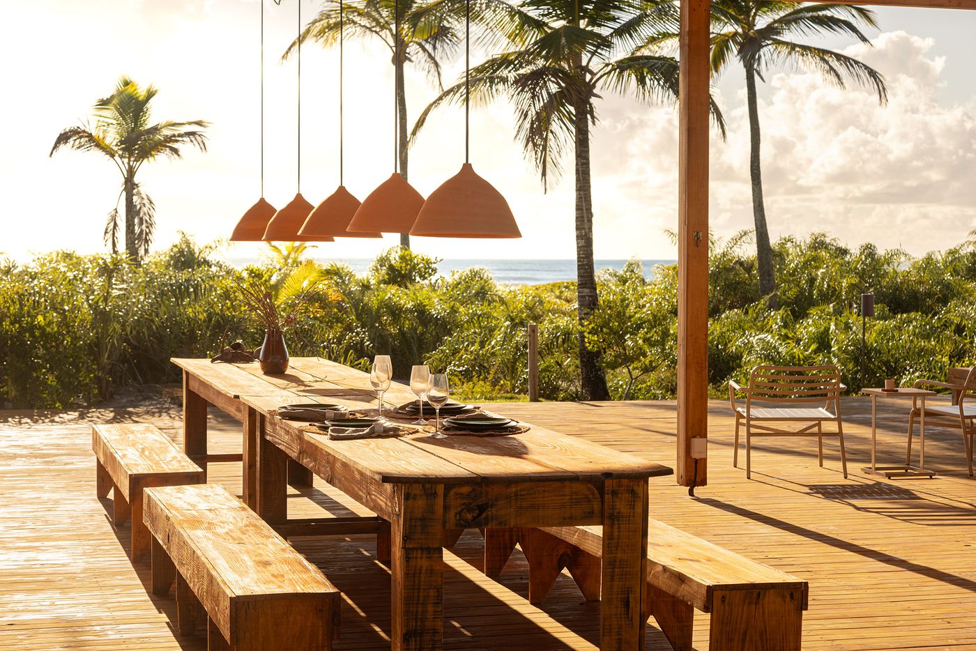 An inviting outdoor dining area on a wooden deck features a long, rustic wooden table with benches, set against a backdrop of palm trees and the ocean at sunset. Ceramic pendant lights hang above, contributing to a warm, evocative atmosphere perfect for shared meals. An inviting outdoor dining area on a wooden deck features a long, rustic wooden table with benches, set against a backdrop of palm trees and the ocean at sunset. Ceramic pendant lights hang above, contributing to a warm, evocative atmosphere perfect for shared meals.
