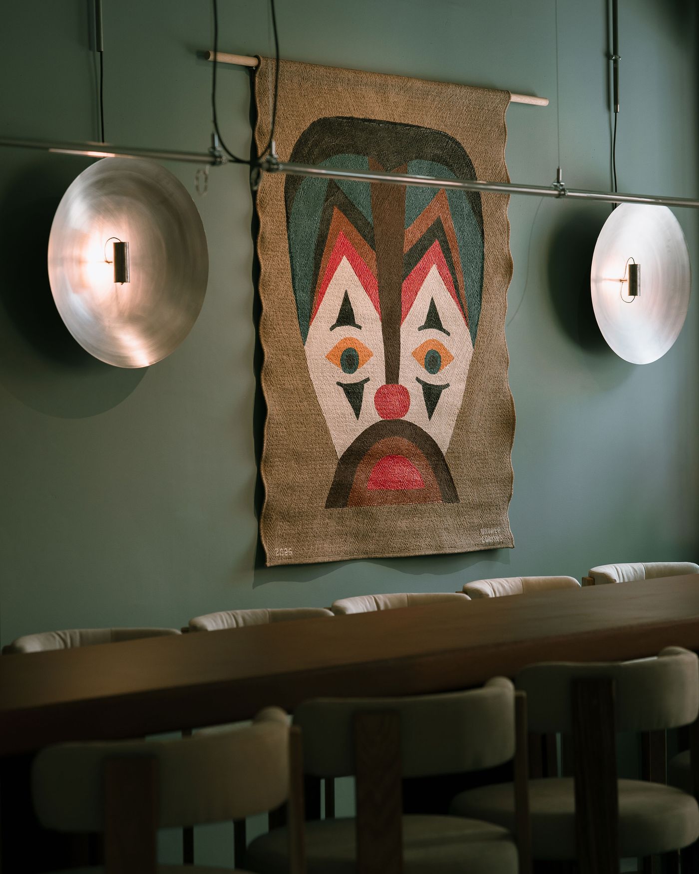 Colorful tribal mask artwork hangs on a teal wall, flanked by modern round light fixtures, with a wooden table and upholstered chairs below.