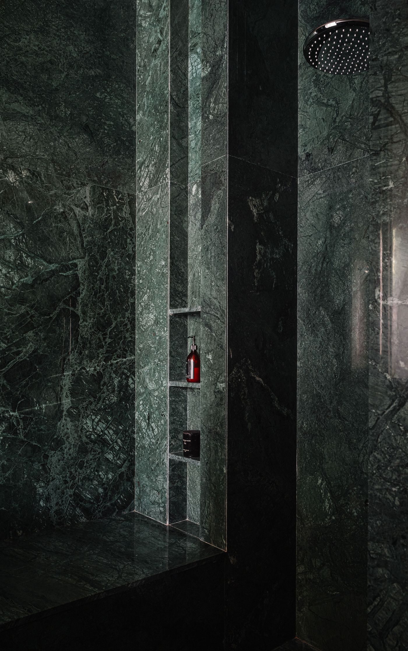 A luxurious shower enclosure entirely clad in dark green marble with prominent white veining, creating a dramatic and immersive experience. Recessed shelves within the marble wall hold small bottles of bath products. A rain showerhead is visible above, emphasizing the seamless and opulent design.
