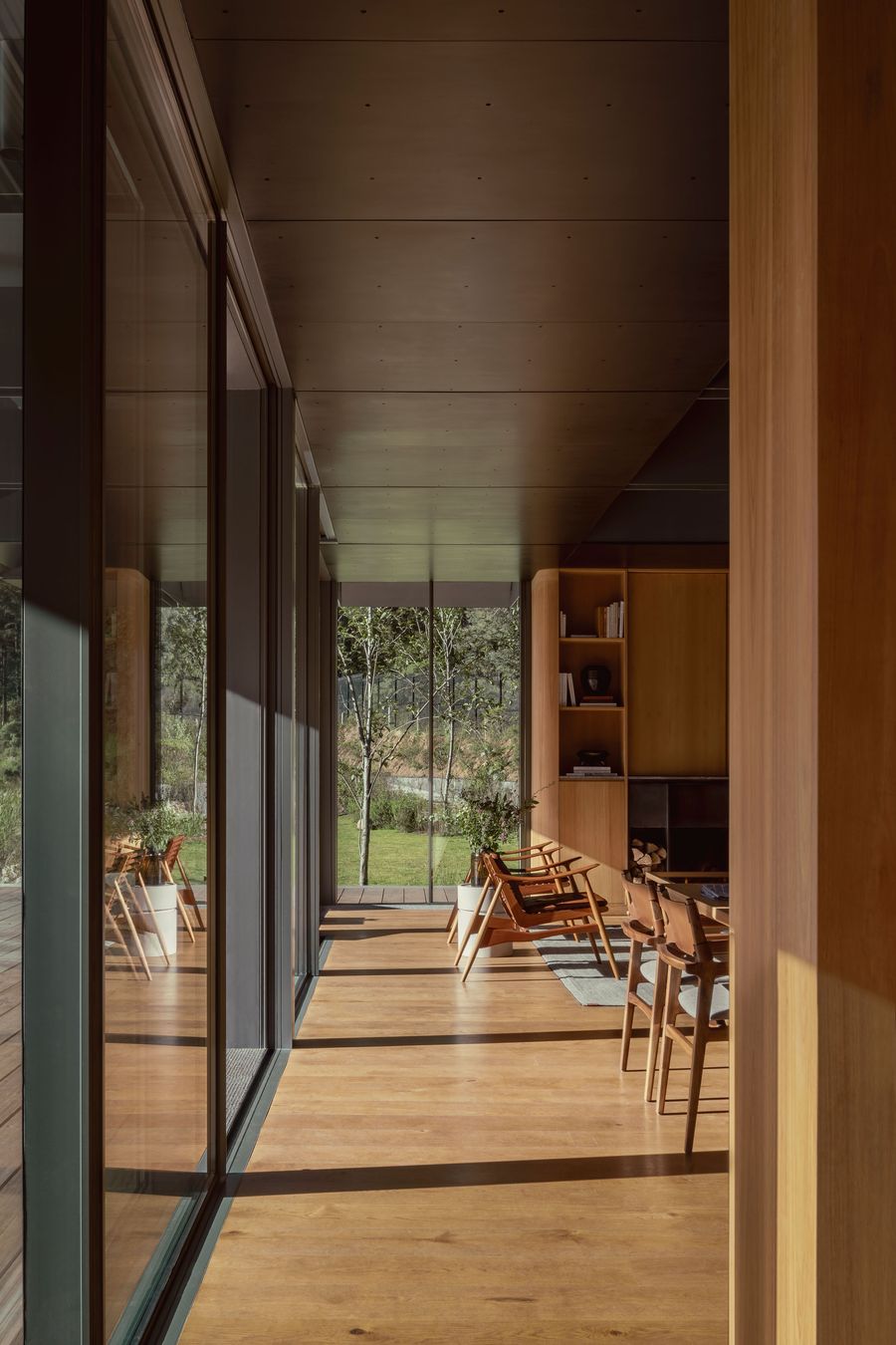 A modern interior hallway with wooden floors, large glass windows, and a view of greenery beyond, featuring cozy seating and shelving.