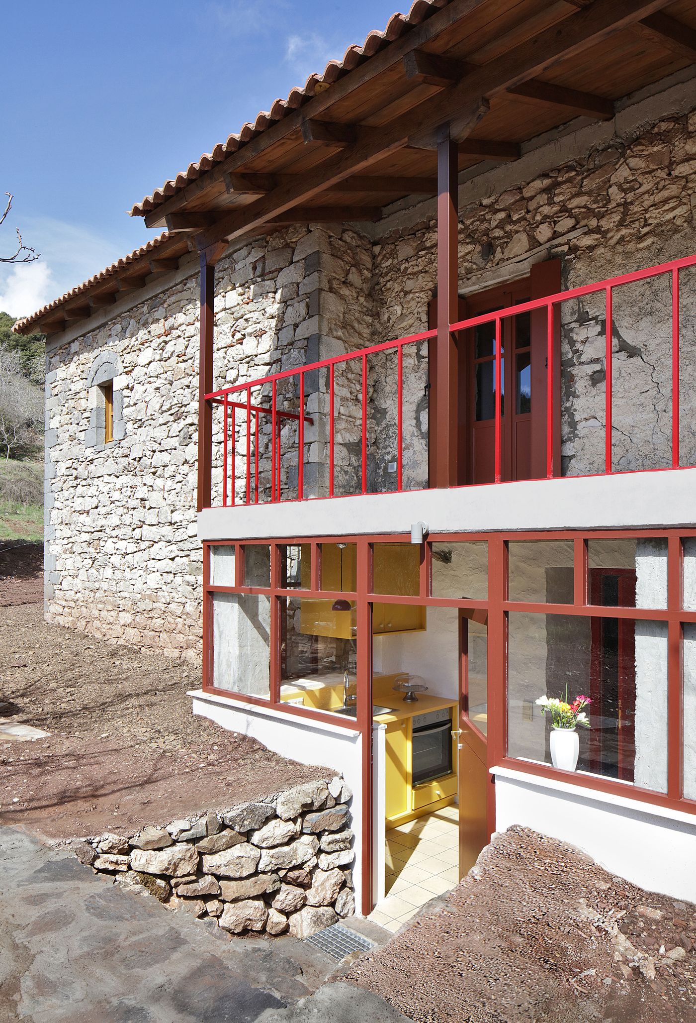 Large-scale glazing opens the renovated interior to natural light and landscape views. The transparent ground floor contrasts with the solid stone upper level, while coral-red framing unifies the contemporary intervention with consistent material language throughout the project.