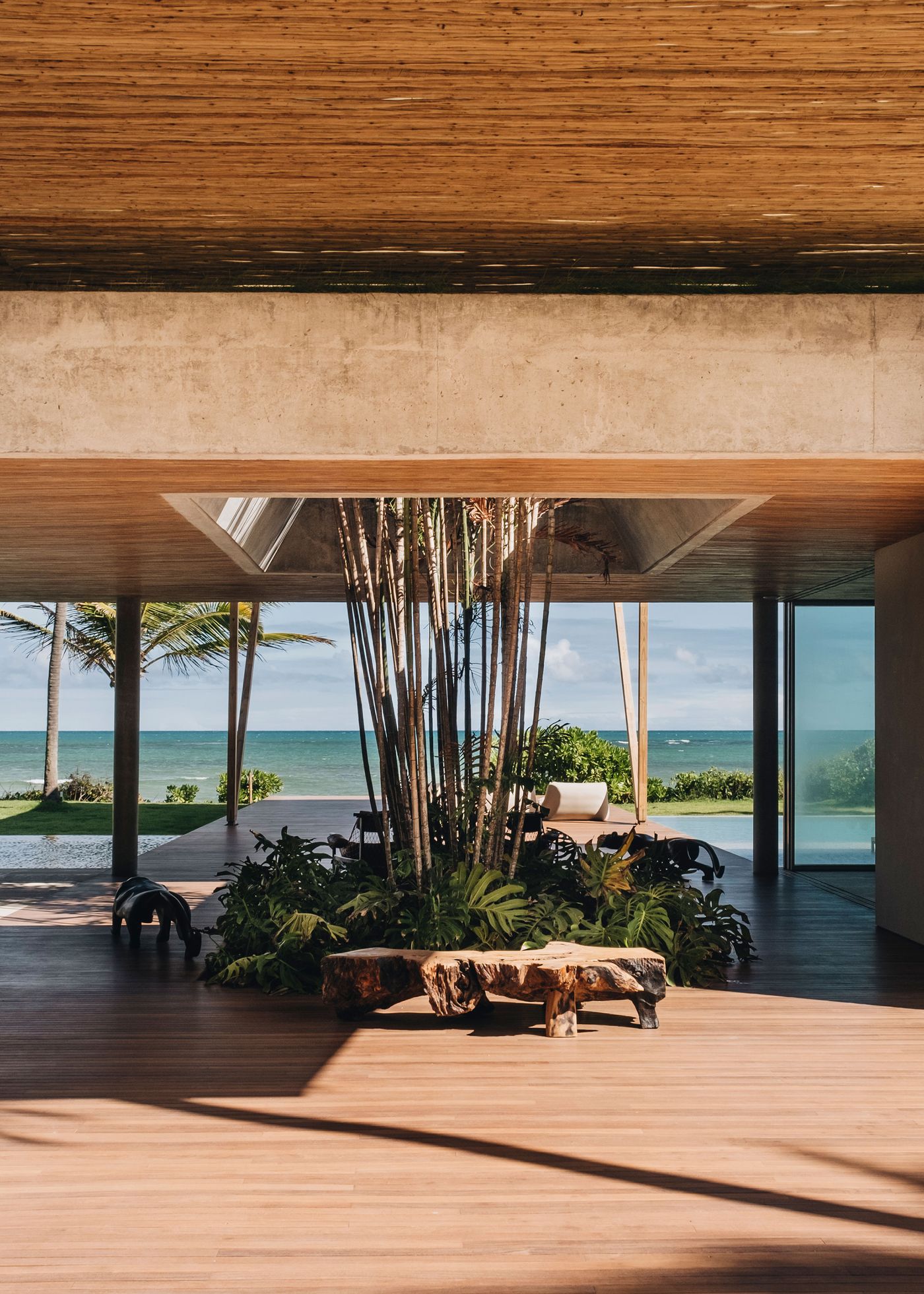 A modern beachfront interior showcasing bamboo ceilings, tropical plants, and views of the ocean through expansive glass walls.