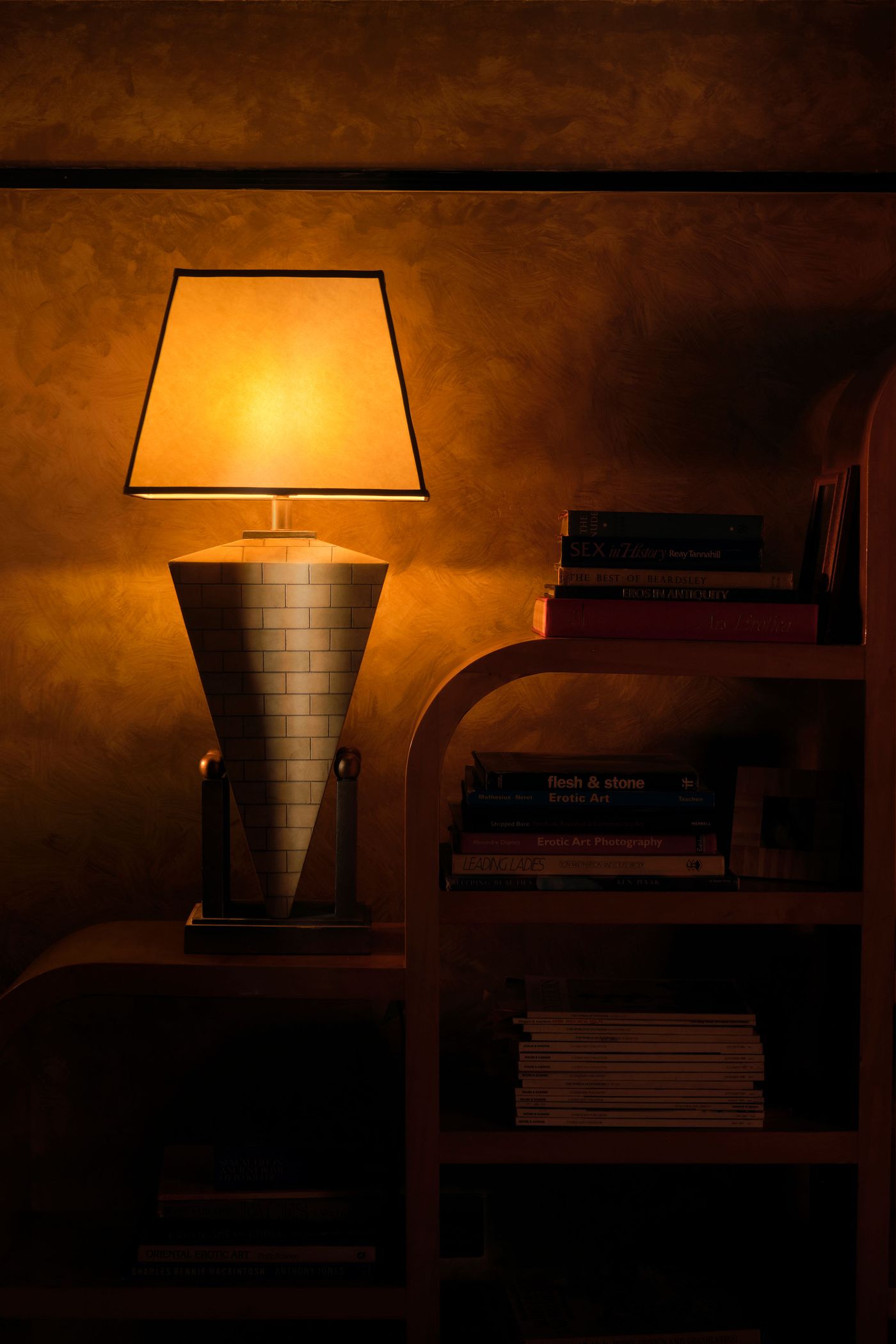 A vintage-retro inspired table lamp with a dark-edged rectangular shade sits on a dark wooden surface, illuminating a stack of books and a curved bookshelf in a warm, inviting glow. The textured, amber-toned wall provides a rich backdrop, contributing to the intimate and sophisticated atmosphere of this London establishment.
