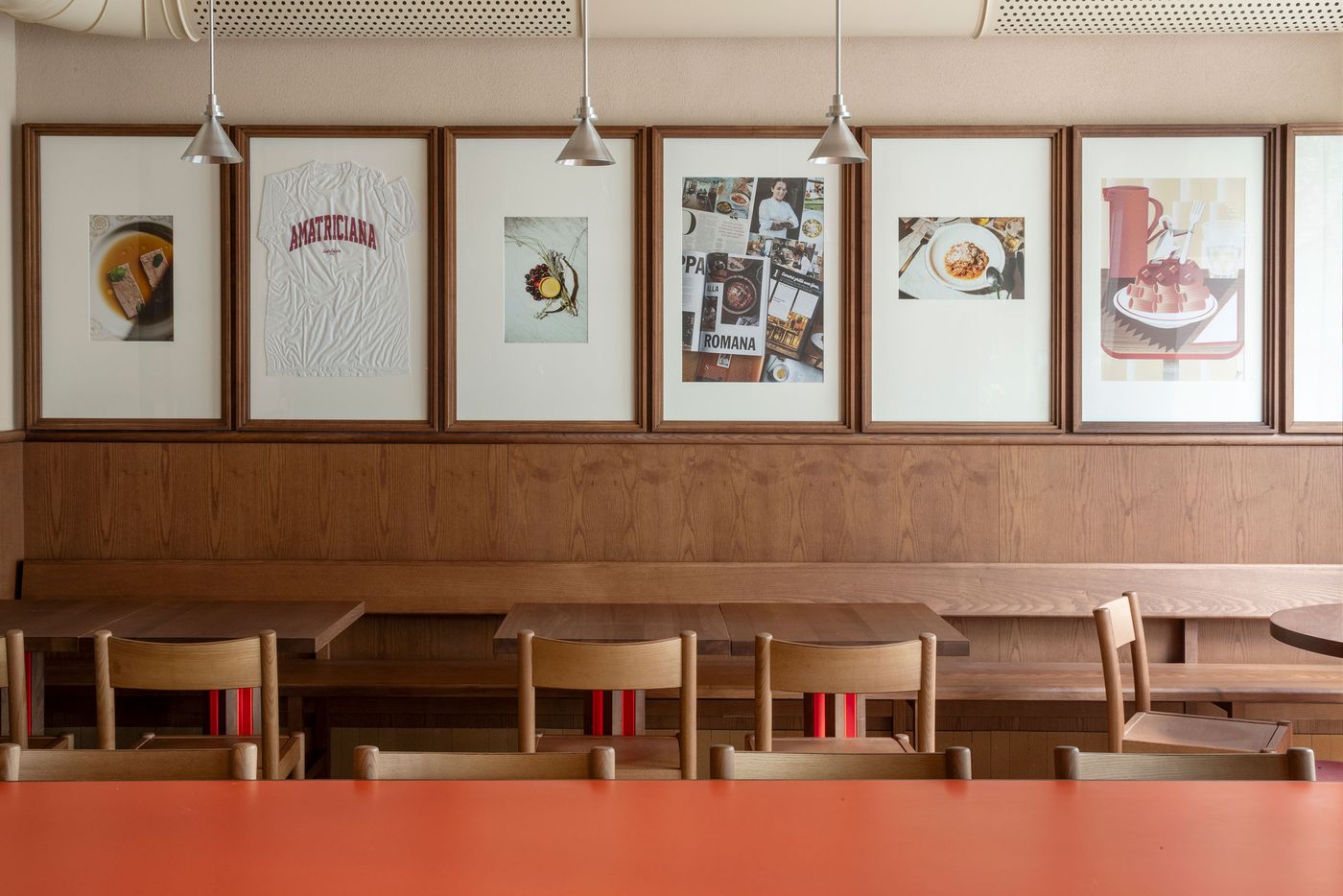 A close-up of Santo Palato's inviting dining area, designed by Naessi Studio, revealing a focus on authentic details. Above the deep wooden banquette, a curated collection of framed artworks, including a graphic of "AMATRICIANA," reflects the restaurant's culinary identity. Industrial pendant lights provide a soft glow, complementing the earthy palette of wood and cream tones. A close-up of Santo Palato's inviting dining area, designed by Naessi Studio, revealing a focus on authentic details. Above the deep wooden banquette, a curated collection of framed artworks, including a graphic of "AMATRICIANA," reflects the restaurant's culinary identity. Industrial pendant lights provide a soft glow, complementing the earthy palette of wood and cream tones.
