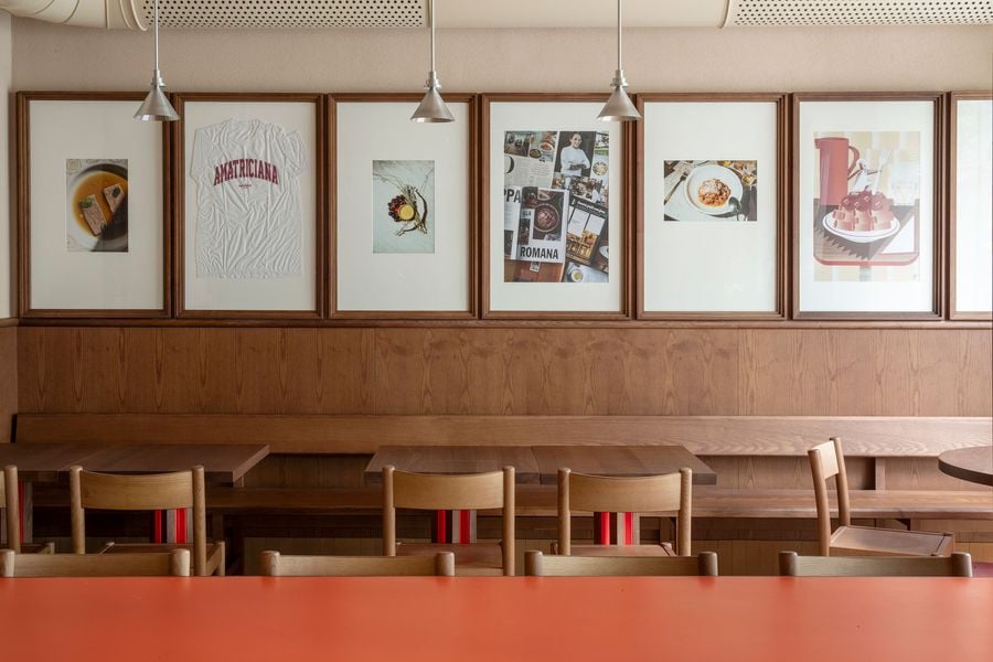 A close-up of Santo Palato's inviting dining area, designed by Naessi Studio, revealing a focus on authentic details. Above the deep wooden banquette, a curated collection of framed artworks, including a graphic of "AMATRICIANA," reflects the restaurant's culinary identity. Industrial pendant lights provide a soft glow, complementing the earthy palette of wood and cream tones.