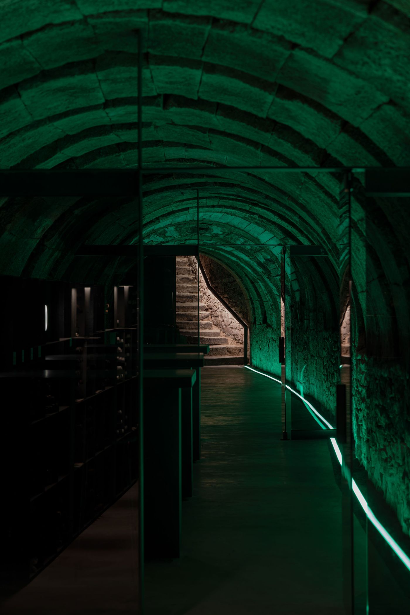 A dimly lit underground corridor with green lighting and stone arches, leading to illuminated stairs in the background.