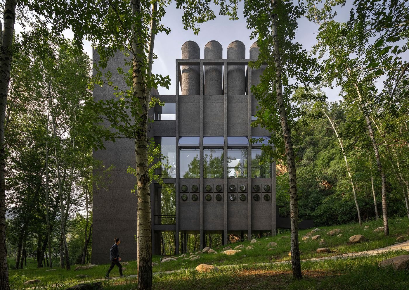 A modern, gray building with cylindrical elements stands among lush greenery, as a person walks along a path nearby. A modern, gray building with cylindrical elements stands among lush greenery, as a person walks along a path nearby.