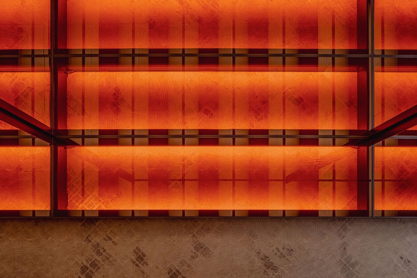 A close view of the basement’s illuminated glass floor reveals layers of orange and red light beneath translucent panels marked by subtle surface textures. Dark structural framing introduces a strict grid, transforming the circulation surface into a luminous architectural element rather than a conventional retail floor.