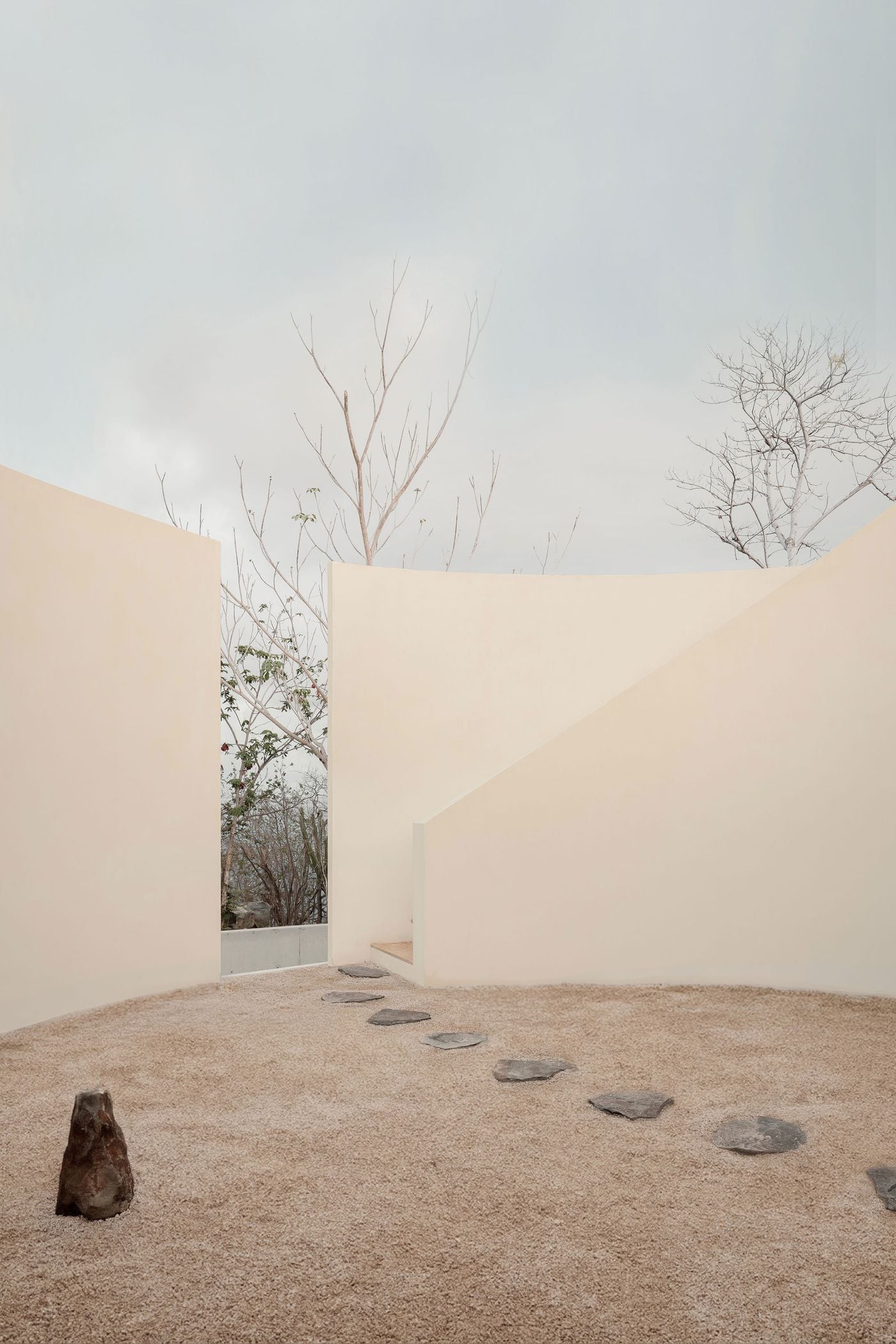 In the round, open-sky courtyard, an arc of slate-grey stones crosses sand-coloured gravel towards a slot in the curving walls, where dry forest briefly breaks through. A single upright rock anchors the foreground, while a raking wall conceals the stair that climbs towards the roof. In the round, open-sky courtyard, an arc of slate-grey stones crosses sand-coloured gravel towards a slot in the curving walls, where dry forest briefly breaks through. A single upright rock anchors the foreground, while a raking wall conceals the stair that climbs towards the roof.