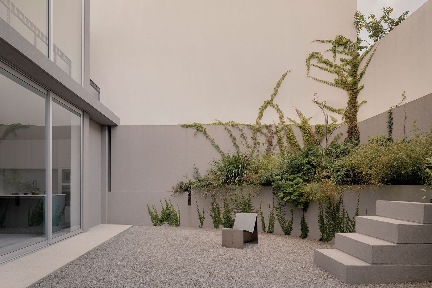 The rear courtyard combines an expanse of pale gravel with dense, layered planting that spills from raised beds and climbs the enclosing walls. Broad concrete steps ascend through the greenery, while a solitary metal chair and the adjacent glazed living room reinforce the garden’s deliberately spare, almost Zen-like character. The rear courtyard combines an expanse of pale gravel with dense, layered planting that spills from raised beds and climbs the enclosing walls. Broad concrete steps ascend through the greenery, while a solitary metal chair and the adjacent glazed living room reinforce the garden’s deliberately spare, almost Zen-like character.
