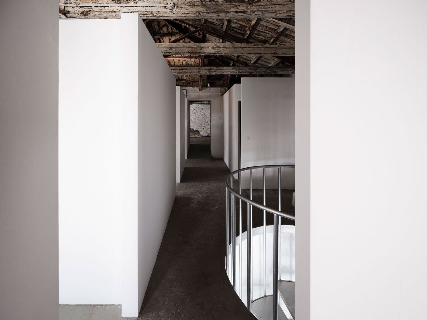 On the upper level, crisp white partitions retained from the building’s later occupation form a sequence of gallery spaces beneath the exposed 150-year-old timber roof. The weathered beams and boards provide a striking counterpoint to the pared-back walls, while the curved metal balustrade marks the spiral staircase below. On the upper level, crisp white partitions retained from the building’s later occupation form a sequence of gallery spaces beneath the exposed 150-year-old timber roof. The weathered beams and boards provide a striking counterpoint to the pared-back walls, while the curved metal balustrade marks the spiral staircase below.