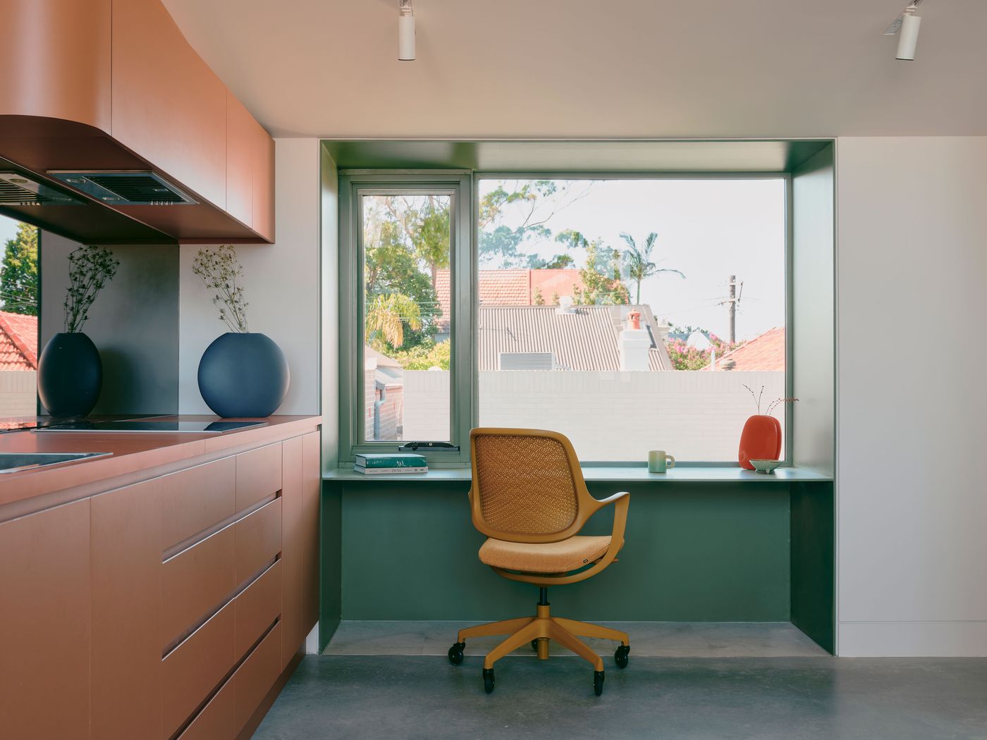 A built-in workspace occupies a deep sage-green window alcove beside the terracotta kitchen, demonstrating how custom joinery maximises Denj Lane’s compact interior. A mustard-toned cane office chair and sculptural ceramics add warmth, while the large window frames neighbouring rooftops and floods the minimalist space with daylight. A built-in workspace occupies a deep sage-green window alcove beside the terracotta kitchen, demonstrating how custom joinery maximises Denj Lane’s compact interior. A mustard-toned cane office chair and sculptural ceramics add warmth, while the large window frames neighbouring rooftops and floods the minimalist space with daylight.
