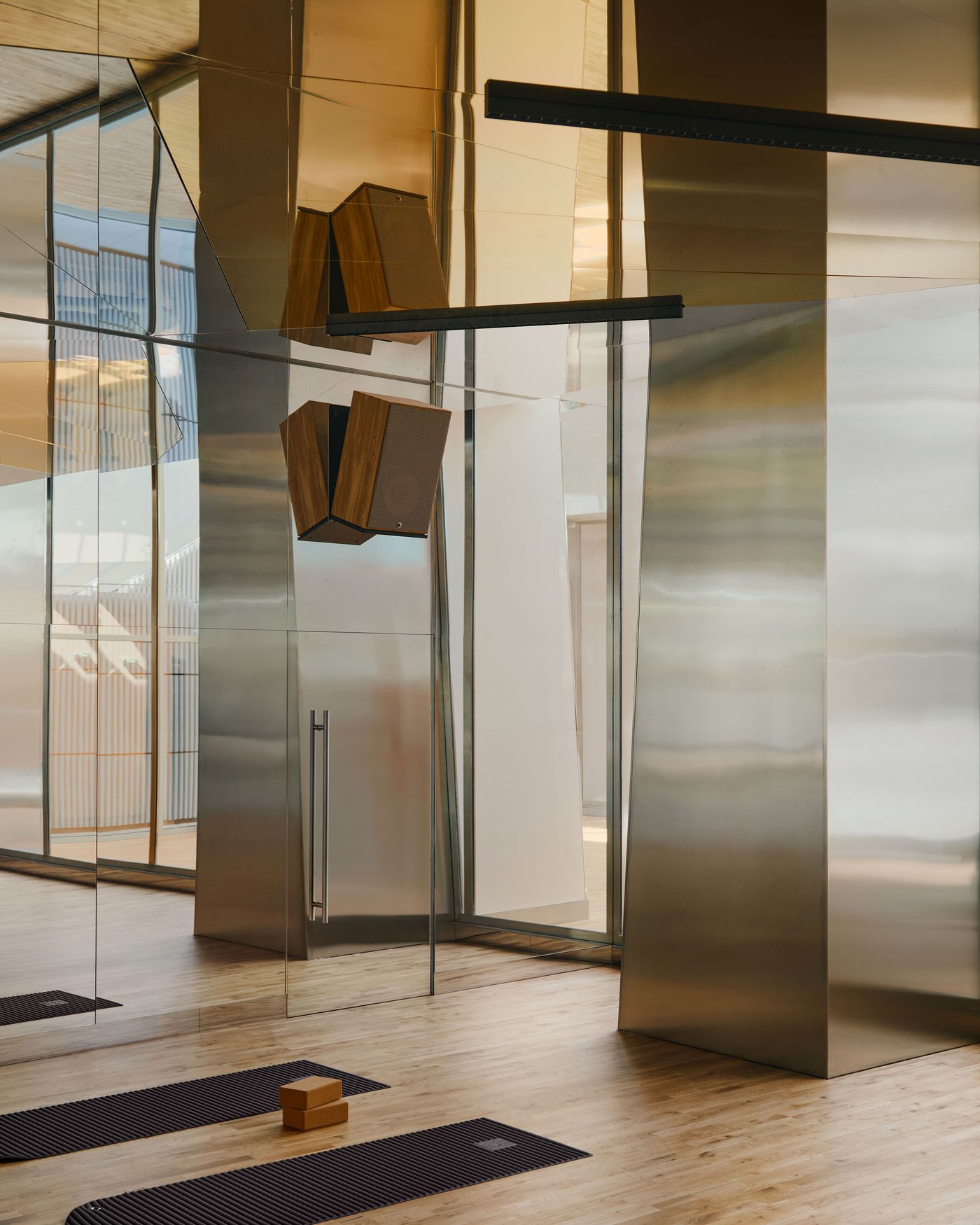 A quieter studio within the Ken Club is defined by timber flooring, mirrored walls and large stainless-steel volumes that catch warm natural light. Minimal black mats and wooden blocks keep the space visually restrained, while suspended linear lights and wall-mounted speakers introduce a sharper, contemporary edge. A quieter studio within the Ken Club is defined by timber flooring, mirrored walls and large stainless-steel volumes that catch warm natural light. Minimal black mats and wooden blocks keep the space visually restrained, while suspended linear lights and wall-mounted speakers introduce a sharper, contemporary edge.