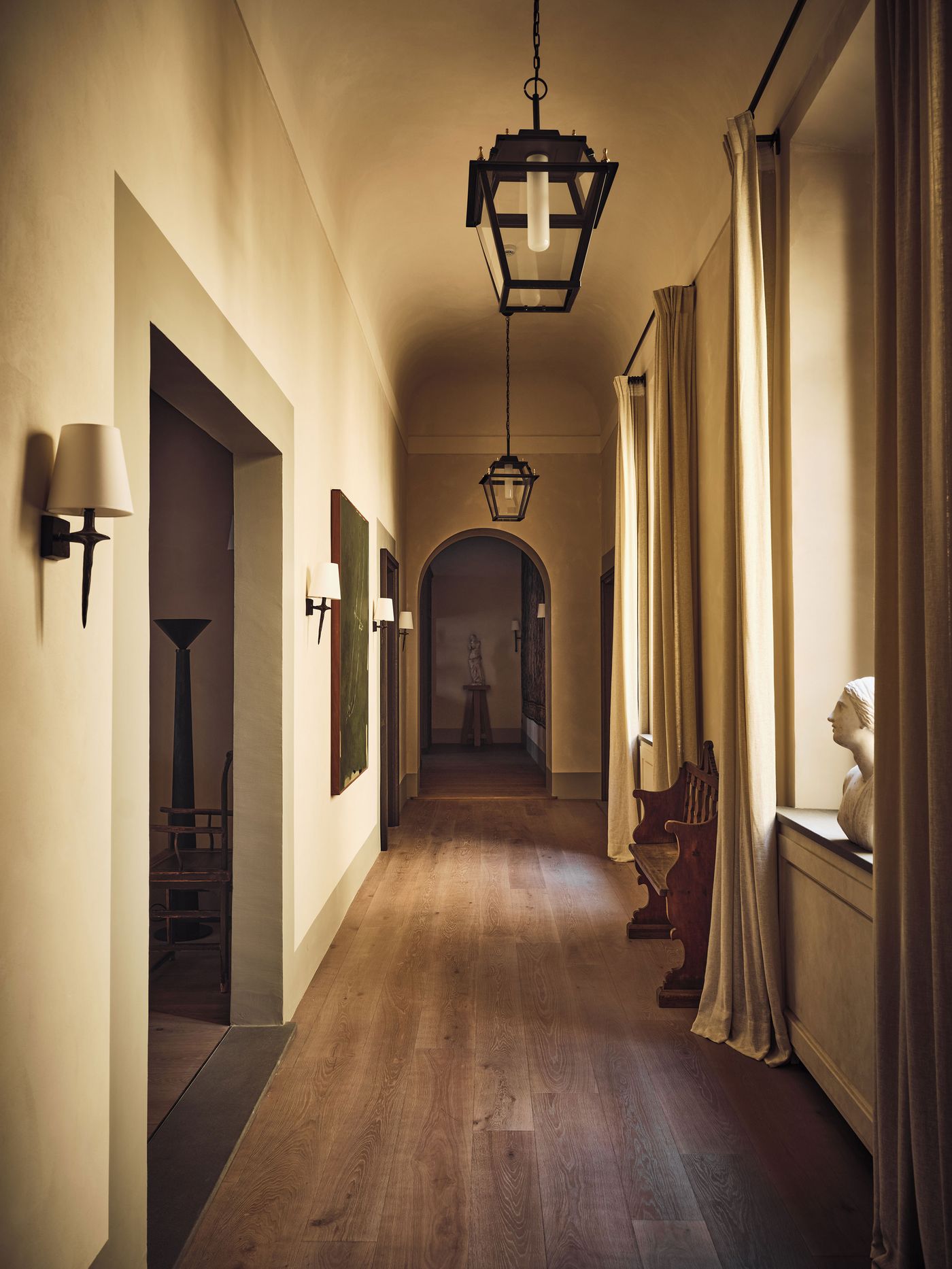 A barrel-vaulted corridor at La Réserve Firenze, lit by glazed iron lanterns and sculptural bronze sconces, leads to an arched opening and a classical statue on a plinth. A marble bust on the deep windowsill, an antique carved bench and a large green abstract canvas punctuate the long run of wide oak boards. A barrel-vaulted corridor at La Réserve Firenze, lit by glazed iron lanterns and sculptural bronze sconces, leads to an arched opening and a classical statue on a plinth. A marble bust on the deep windowsill, an antique carved bench and a large green abstract canvas punctuate the long run of wide oak boards.