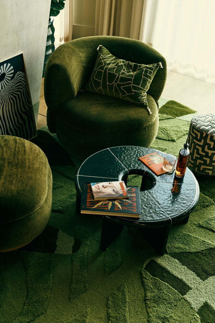 A close-up of the ground-floor lounge highlights its tactile layering of moss-green upholstery, sculptural furniture and a deeply textured geometric rug. A glossy black coffee table, patterned textiles and graphic artwork sharpen the retro palette, while filtered daylight gives this intimate seating zone a relaxed, residential character. A close-up of the ground-floor lounge highlights its tactile layering of moss-green upholstery, sculptural furniture and a deeply textured geometric rug. A glossy black coffee table, patterned textiles and graphic artwork sharpen the retro palette, while filtered daylight gives this intimate seating zone a relaxed, residential character.