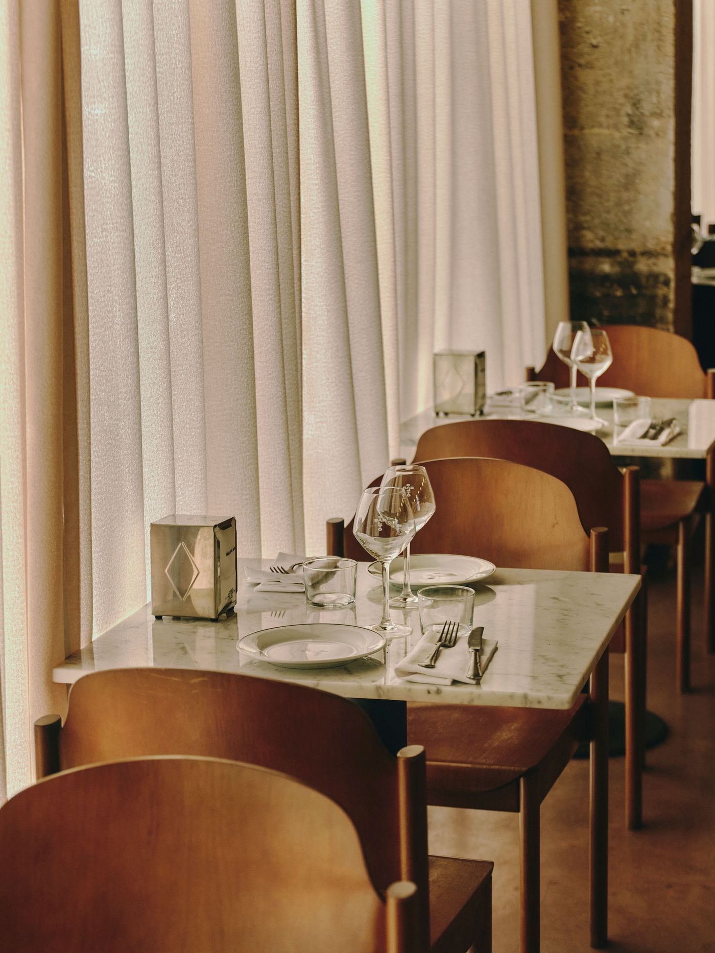 Sheer curtains filter the daylight along the window tables at SaltoBar in Paris, casting a soft, peachy glow over white marble tops. Curved plywood chairs in warm wood tones sit beside steel napkin dispensers with an engraved diamond motif. Wine glasses etched with the SaltoBar name complete the table settings. Sheer curtains filter the daylight along the window tables at SaltoBar in Paris, casting a soft, peachy glow over white marble tops. Curved plywood chairs in warm wood tones sit beside steel napkin dispensers with an engraved diamond motif. Wine glasses etched with the SaltoBar name complete the table settings.