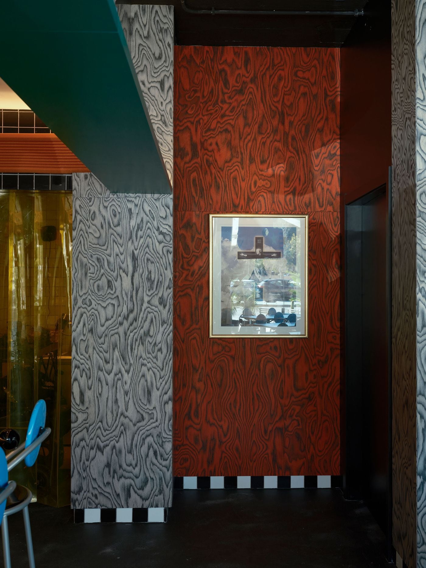 At Seth & Manni's, wood-grey veneer meets a rust-red panel of the same swirling figure, set off by a gilt-framed photographic artwork depicting a crucifixion. A checkerboard skirting runs along the base. An amber strip curtain glows at the left edge, completing a study in contrasting veneers. At Seth & Manni's, wood-grey veneer meets a rust-red panel of the same swirling figure, set off by a gilt-framed photographic artwork depicting a crucifixion. A checkerboard skirting runs along the base. An amber strip curtain glows at the left edge, completing a study in contrasting veneers.