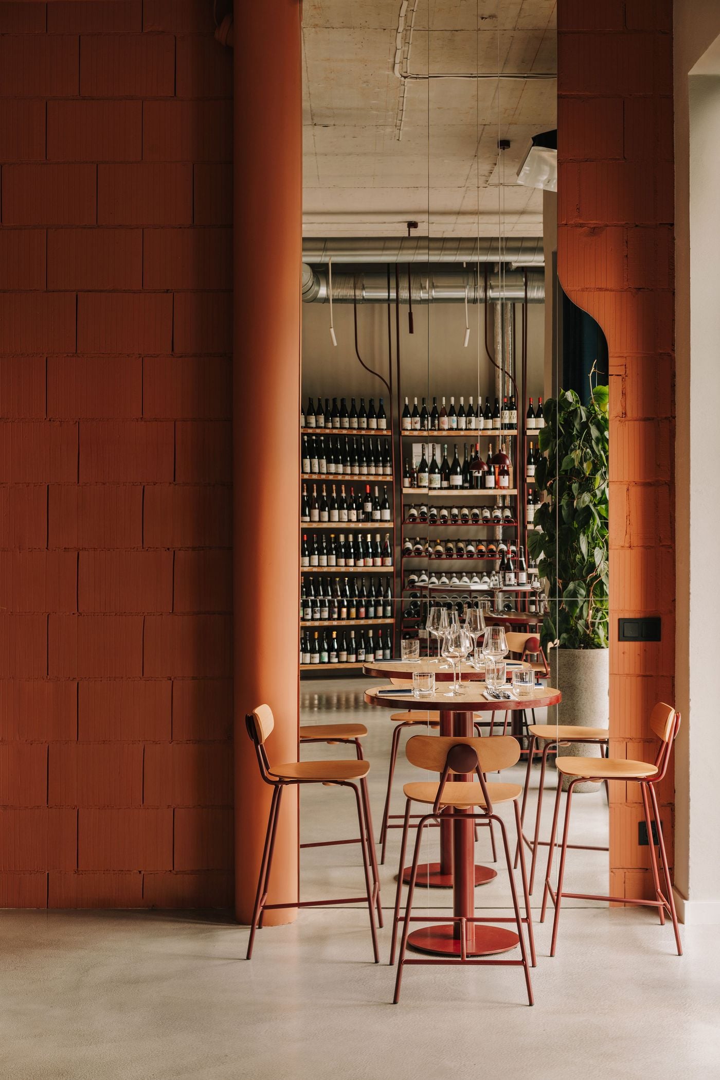 A floor-to-ceiling mirror framed by terracotta-coloured masonry reflects WIN wine bar’s ground-floor retail area, with its neatly arranged wine displays and exposed ventilation ducts. In the foreground, two round timber-topped tables and burgundy-framed chairs create an intimate seating arrangement, while the mirror adds depth and a sense of spatial continuity to the industrial-inspired interior. A floor-to-ceiling mirror framed by terracotta-coloured masonry reflects WIN wine bar’s ground-floor retail area, with its neatly arranged wine displays and exposed ventilation ducts. In the foreground, two round timber-topped tables and burgundy-framed chairs create an intimate seating arrangement, while the mirror adds depth and a sense of spatial continuity to the industrial-inspired interior.