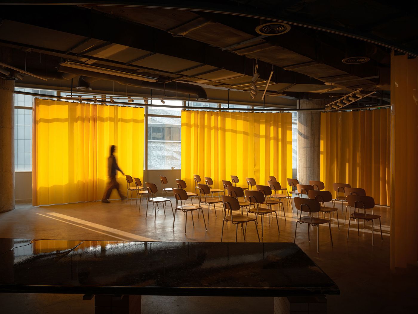 A striking image of 889 GLO Art Space where two large, luminous yellow curtains divide the expansive area, creating a warm, inviting glow. Scattered wooden chairs are arranged on the polished concrete floor, suggesting an event space. A solitary figure walks across the sun-drenched floor, emphasizing the vastness and the atmospheric quality of the industrial interior. A striking image of 889 GLO Art Space where two large, luminous yellow curtains divide the expansive area, creating a warm, inviting glow. Scattered wooden chairs are arranged on the polished concrete floor, suggesting an event space. A solitary figure walks across the sun-drenched floor, emphasizing the vastness and the atmospheric quality of the industrial interior.