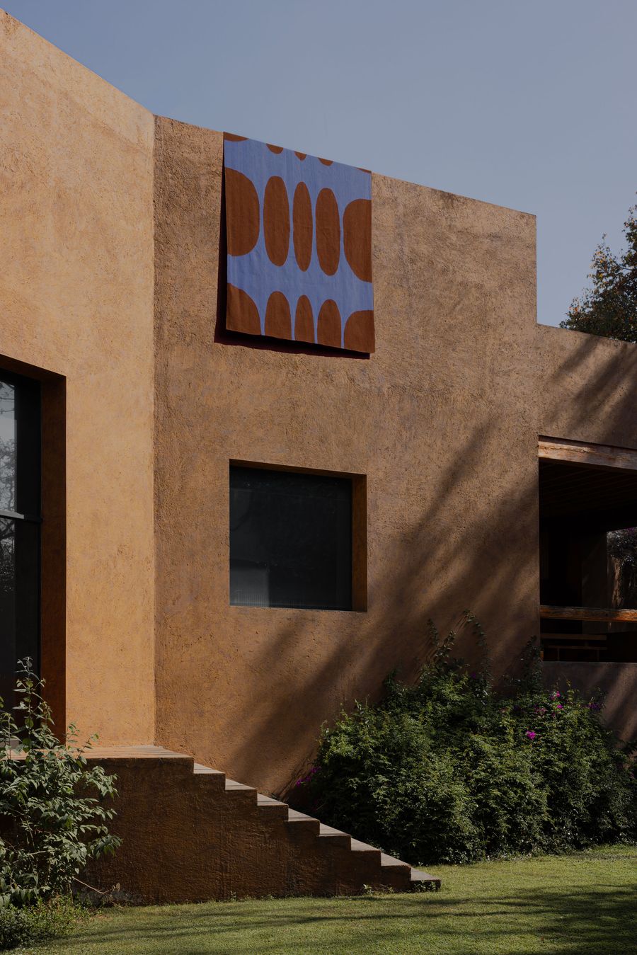 A modern, orange-toned building features a patterned textile hanging on the wall, surrounded by greenery and a clear blue sky.