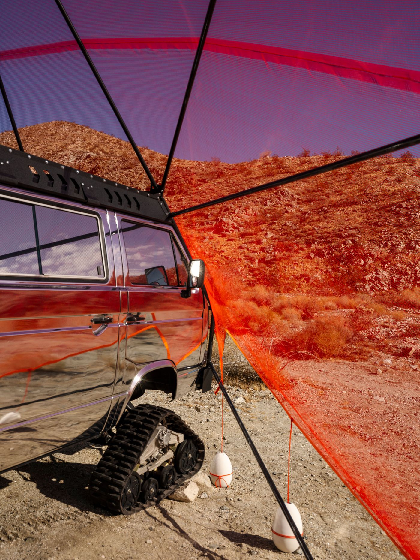 A reflective van with a tank tread, sheltered by a bright orange mesh covering in a rocky desert landscape.