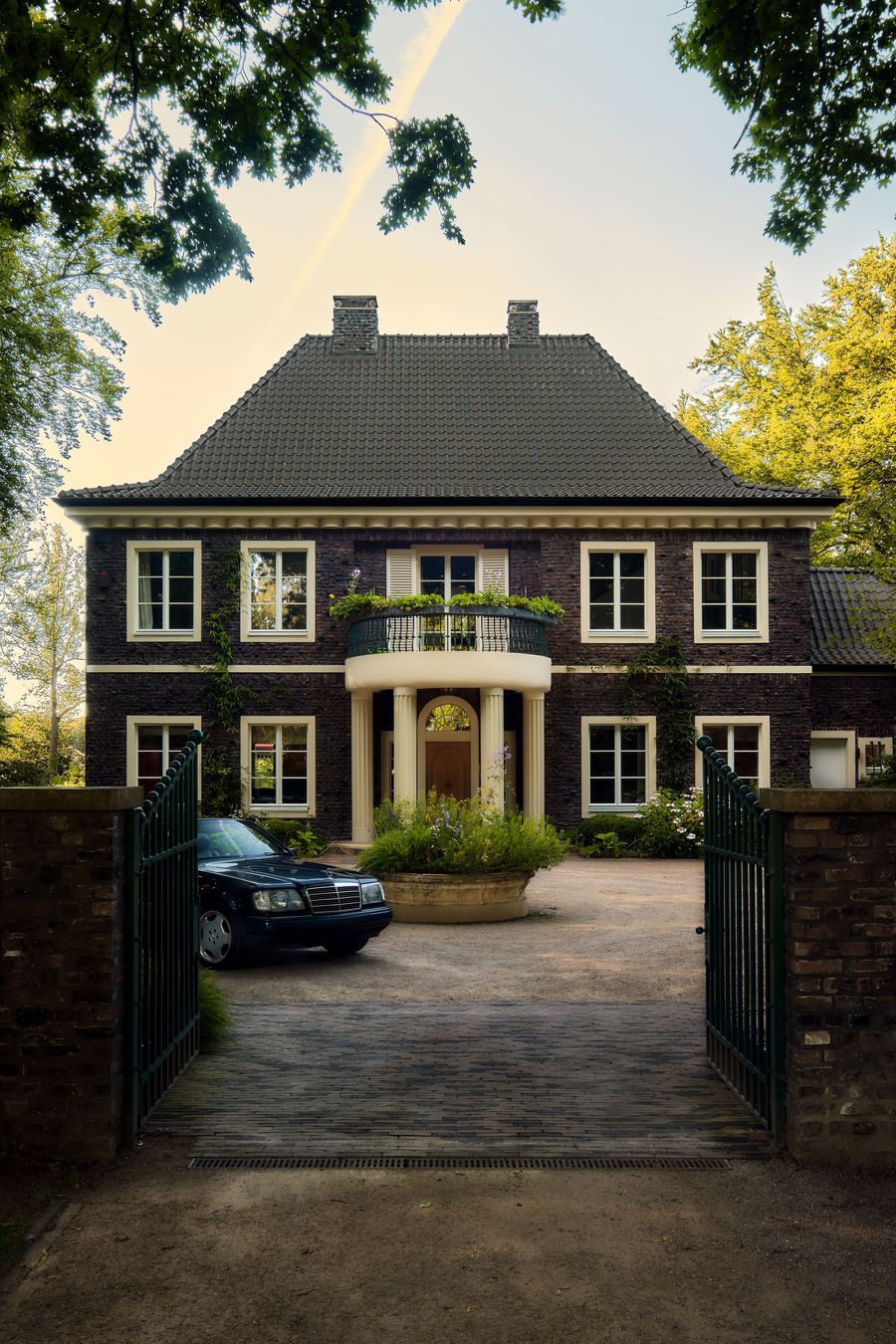 A stately brick house with a front porch, balcony, and lush greenery, framed by trees and a black car parked in the driveway. A stately brick house with a front porch, balcony, and lush greenery, framed by trees and a black car parked in the driveway.