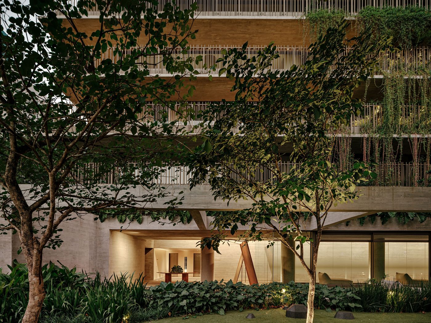 An illuminated evening view of the building's integrated landscape and brutalist architecture. The ground floor's warm interior glow contrasts with the cool tones of the concrete and the verdant, subtly lit garden, emphasizing the seamless transition between built form and natural environment. An illuminated evening view of the building's integrated landscape and brutalist architecture. The ground floor's warm interior glow contrasts with the cool tones of the concrete and the verdant, subtly lit garden, emphasizing the seamless transition between built form and natural environment.