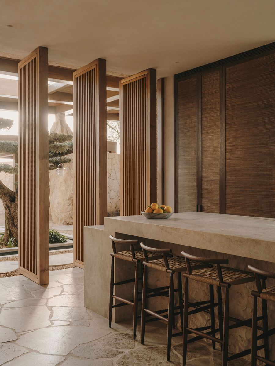 A stylish kitchen with a stone bar, wooden stools, and slatted wooden doors, illuminated by soft natural light and featuring a bowl of lemons. A stylish kitchen with a stone bar, wooden stools, and slatted wooden doors, illuminated by soft natural light and featuring a bowl of lemons.
