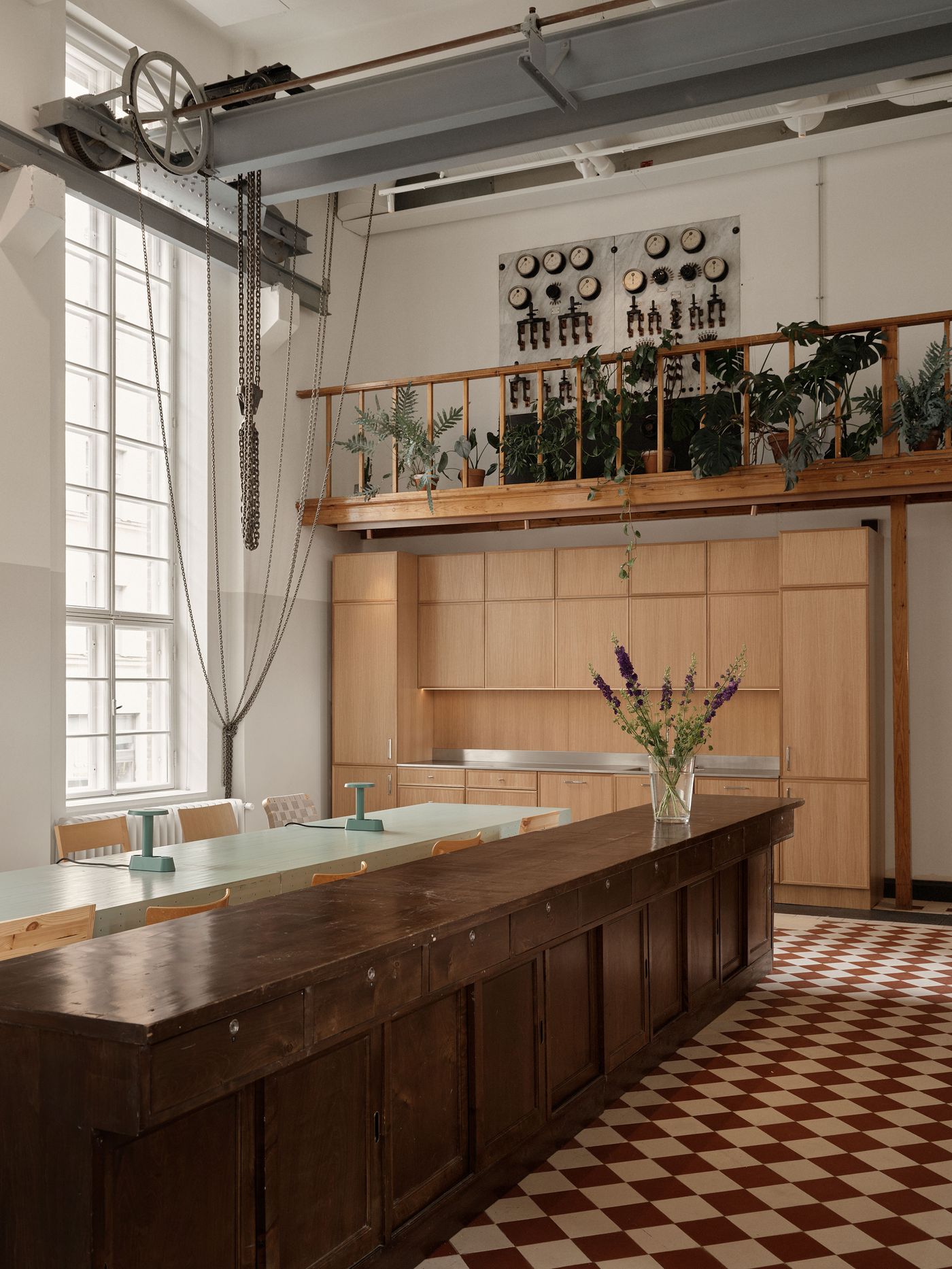 A spacious office kitchen area features a long, light blue table and light wood cabinetry, contrasted with a dark wooden counter. An industrial overhead crane and lush plants on an upper balcony add a unique touch to the vibrant red and white checkered floor space. A spacious office kitchen area features a long, light blue table and light wood cabinetry, contrasted with a dark wooden counter. An industrial overhead crane and lush plants on an upper balcony add a unique touch to the vibrant red and white checkered floor space.