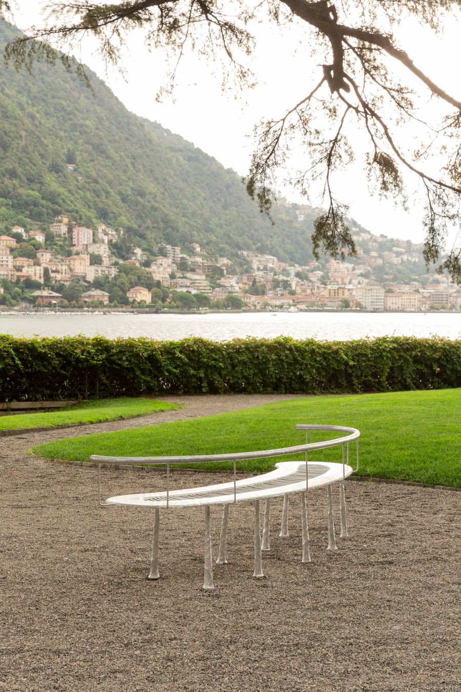 A gracefully curved white metal bench with slender legs is set on a gravel path in a garden overlooking a lake. The organic shape of the bench, with its simple railing, complements the natural curves of the landscape, including a lush green lawn and hedge. The background reveals the picturesque town of Lake Como nestled on a mountainside, showcasing a harmonious blend of modern, minimalist design and natural scenery.