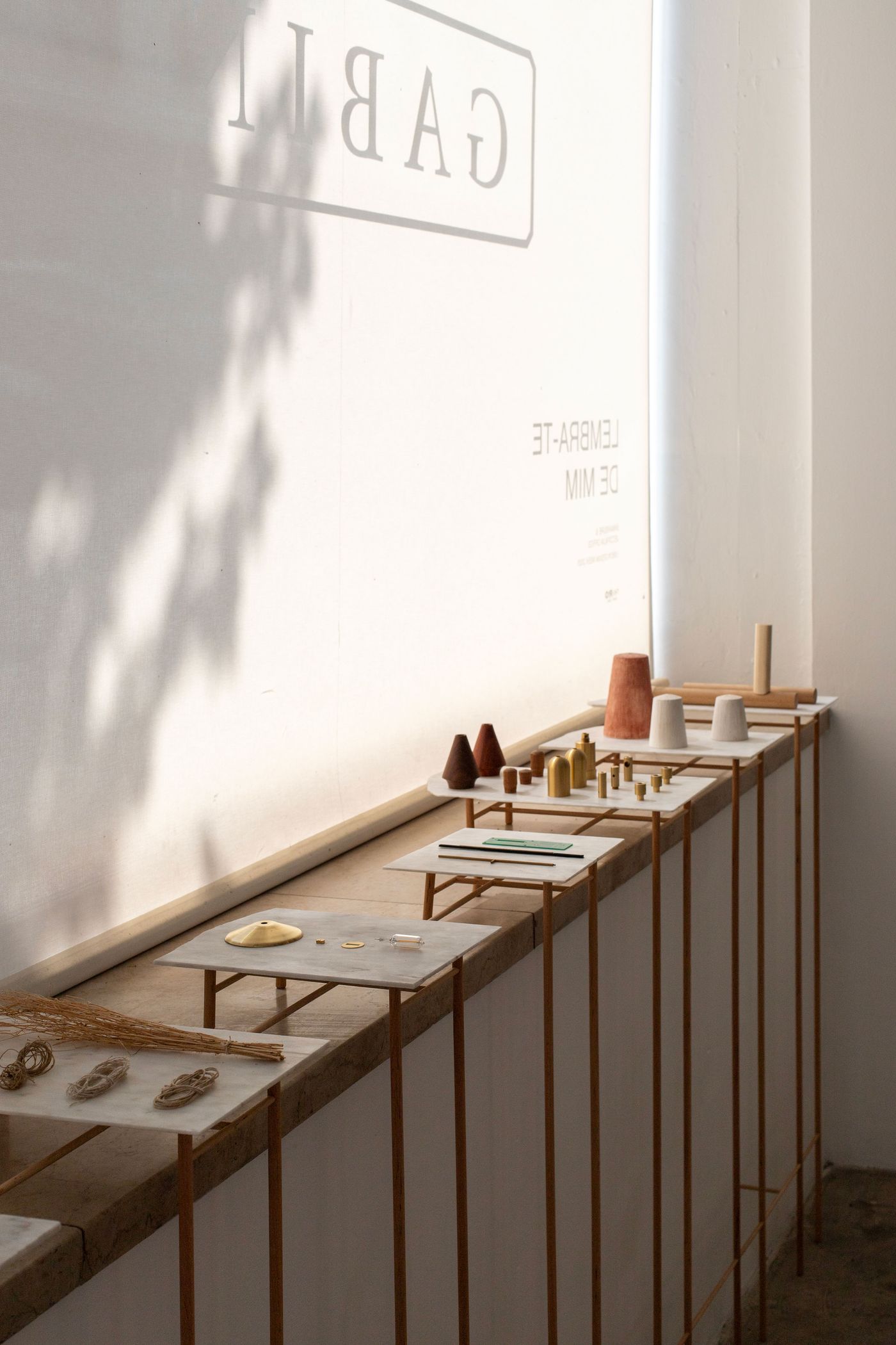 Minimalist wooden display tables create a rhythmic procession along white walls, bathed in gentle natural light. The Scandinavian-inspired arrangement features curated ceramic, metallic and wooden objects, with soft shadows adding depth to this serene gallery-like space celebrating craft and contemplative design.