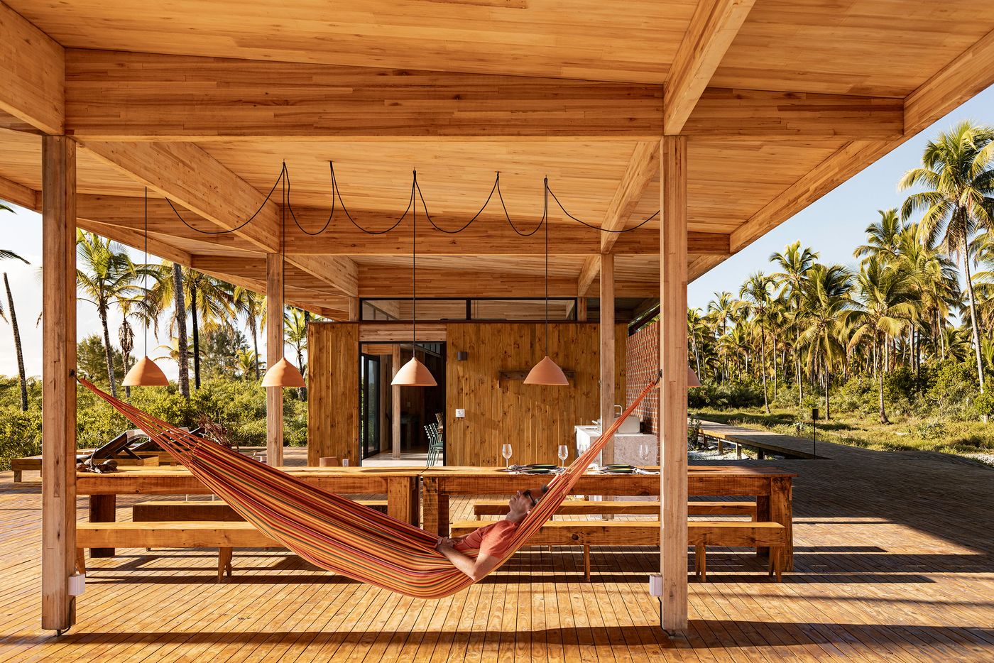 A person relaxes in a striped hammock under a broad wooden roof, part of a modular structure integrated with the tropical landscape. The open-air design, raw wood textures, and the surrounding palm trees create a casual, biophilic atmosphere, inviting outdoor living. A person relaxes in a striped hammock under a broad wooden roof, part of a modular structure integrated with the tropical landscape. The open-air design, raw wood textures, and the surrounding palm trees create a casual, biophilic atmosphere, inviting outdoor living.