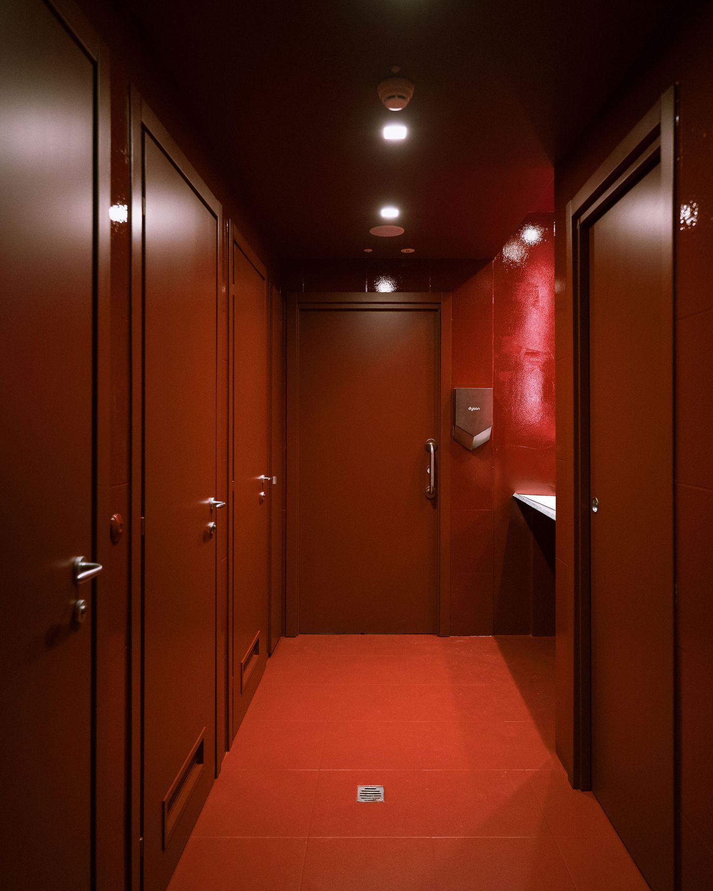 A narrow hallway of a restroom featuring red walls and doors, with bright ceiling lights and a hand dryer visible.