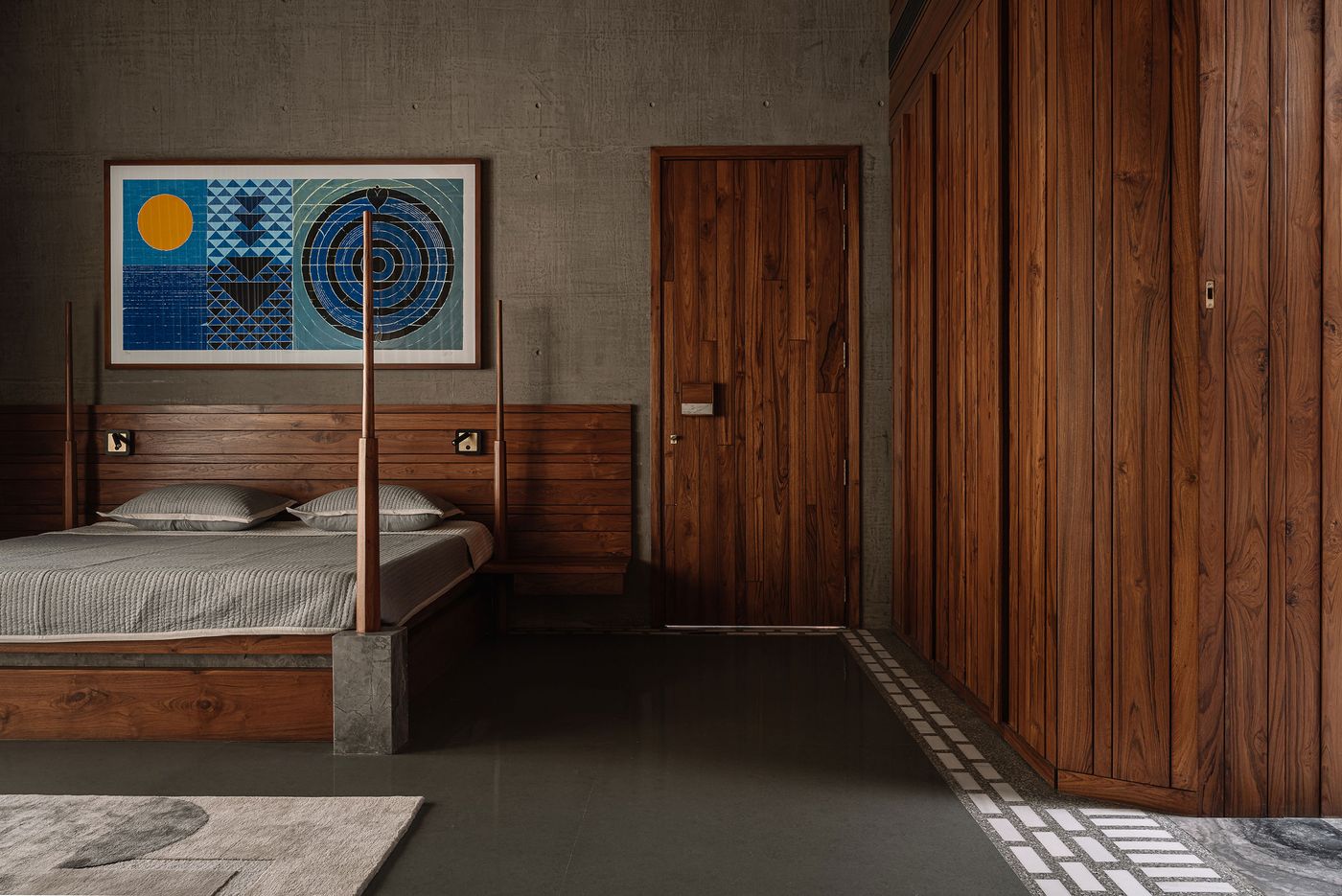 A bedroom defined by raw concrete walls and rich wooden elements, including a low-profile bed and floor-to-ceiling wardrobes. A vibrant geometric artwork with blue, yellow, and black tones adds a focal point. The dark floor with subtle white tiling patterns, combined with the warm wood, creates a balanced, sophisticated aesthetic with a modern edge.