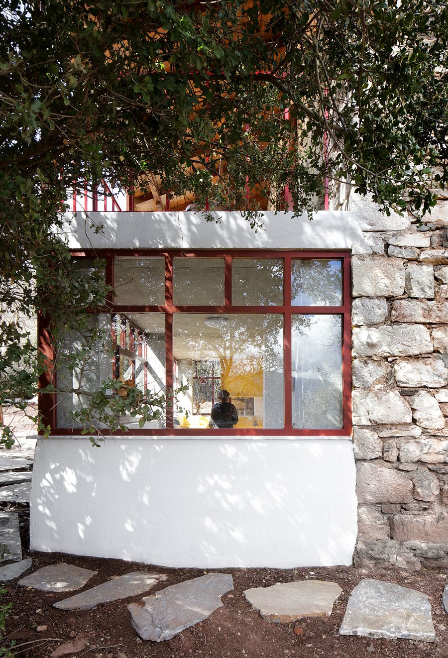 Dappled Mediterranean light filters through ancient olive branches onto contemporary coral-red steel glazing. The biophilic design seamlessly merges interior and exterior realms, where natural stone pathways and verdant canopy create an immersive dialogue between built heritage and landscape.
