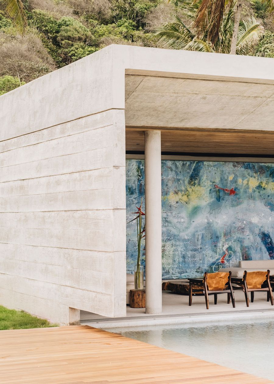 Modern concrete house with a vibrant blue mural, surrounded by greenery and a wooden deck by a tranquil pool.
