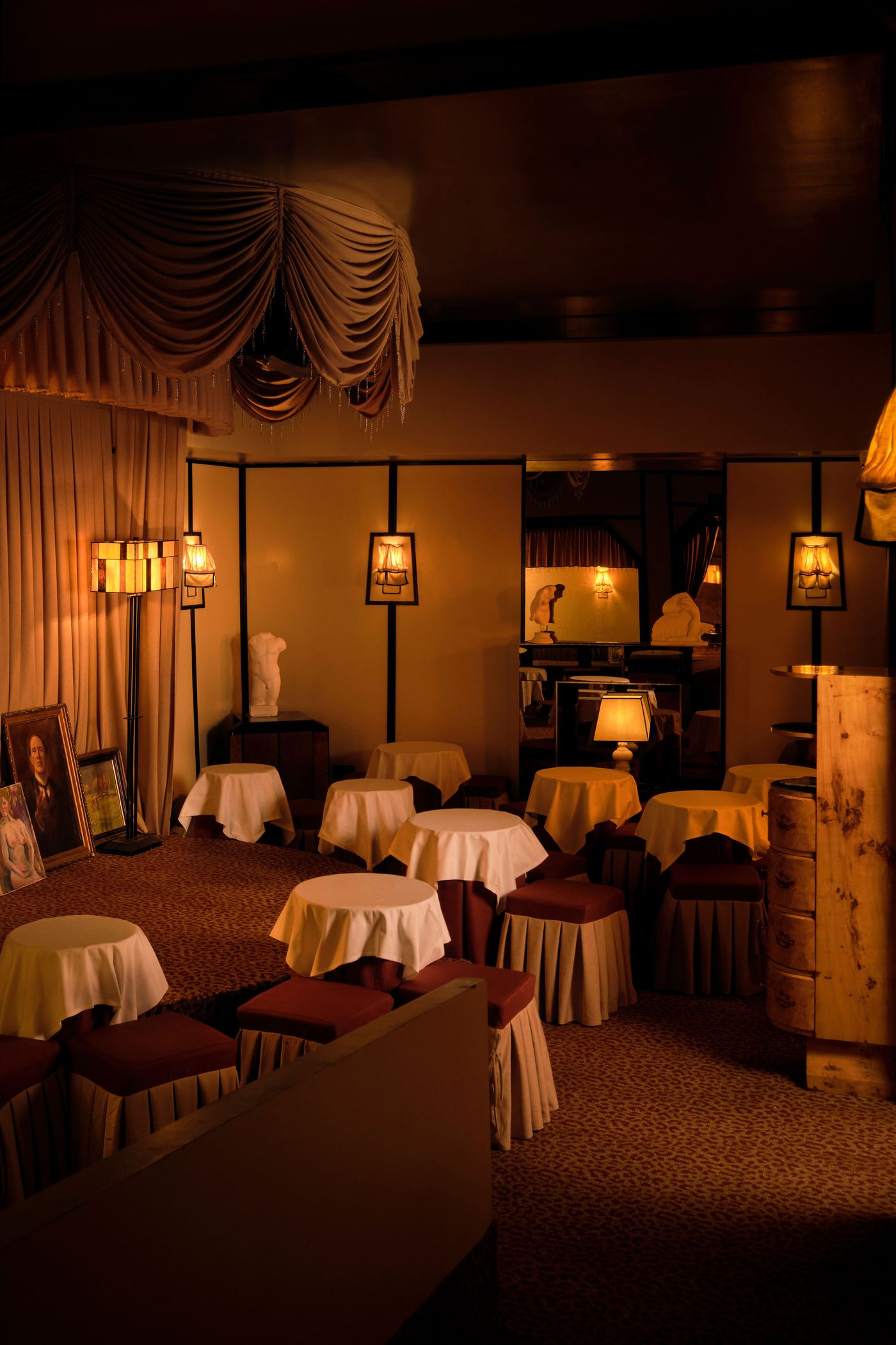 A dimly lit, eclectic lounge area in The Roses of Elagabalus, featuring small, white-draped round tables surrounded by plush, skirted stools in deep red. Ornate lighting, including a floor lamp and unique framed wall sconces, casts warm glows. A classical bust, framed portrait, and richly patterned carpet contribute to the vintage-retro aesthetic and intimate atmosphere. A dimly lit, eclectic lounge area in The Roses of Elagabalus, featuring small, white-draped round tables surrounded by plush, skirted stools in deep red. Ornate lighting, including a floor lamp and unique framed wall sconces, casts warm glows. A classical bust, framed portrait, and richly patterned carpet contribute to the vintage-retro aesthetic and intimate atmosphere.