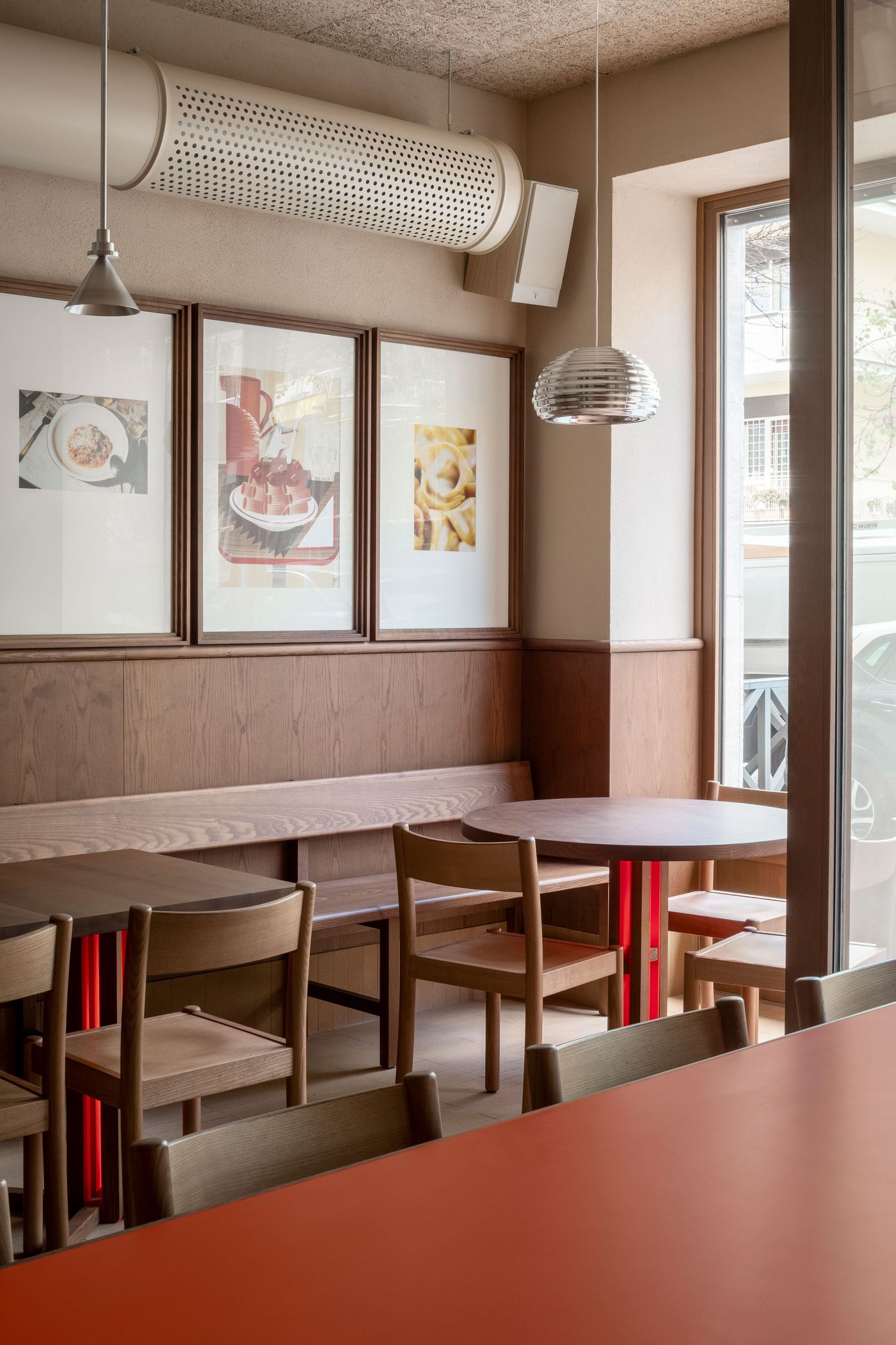 A bustling corner of Santo Palato, designed by Naessi Studio, with a curved wooden banquette providing flexible seating. Warm wood tones dominate the space, contrasted by a bold orange communal table in the foreground. Industrial pendant lights and framed food art contribute to the trattoria's authentic yet modern Roman atmosphere. A bustling corner of Santo Palato, designed by Naessi Studio, with a curved wooden banquette providing flexible seating. Warm wood tones dominate the space, contrasted by a bold orange communal table in the foreground. Industrial pendant lights and framed food art contribute to the trattoria's authentic yet modern Roman atmosphere.