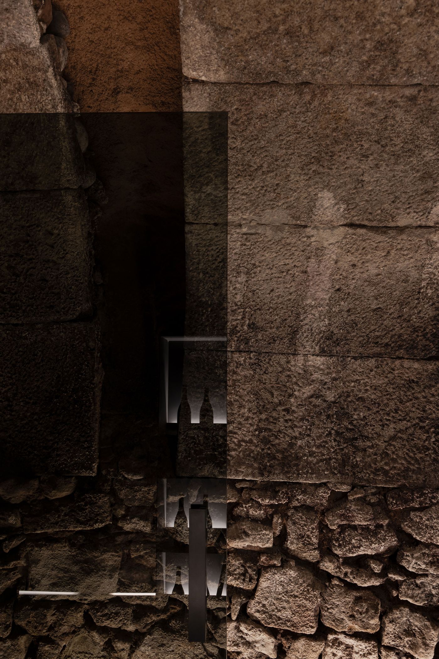 An abstract view of textured stone walls, with a dark overlay and ambient light revealing silhouettes of bottles in the background. An abstract view of textured stone walls, with a dark overlay and ambient light revealing silhouettes of bottles in the background.