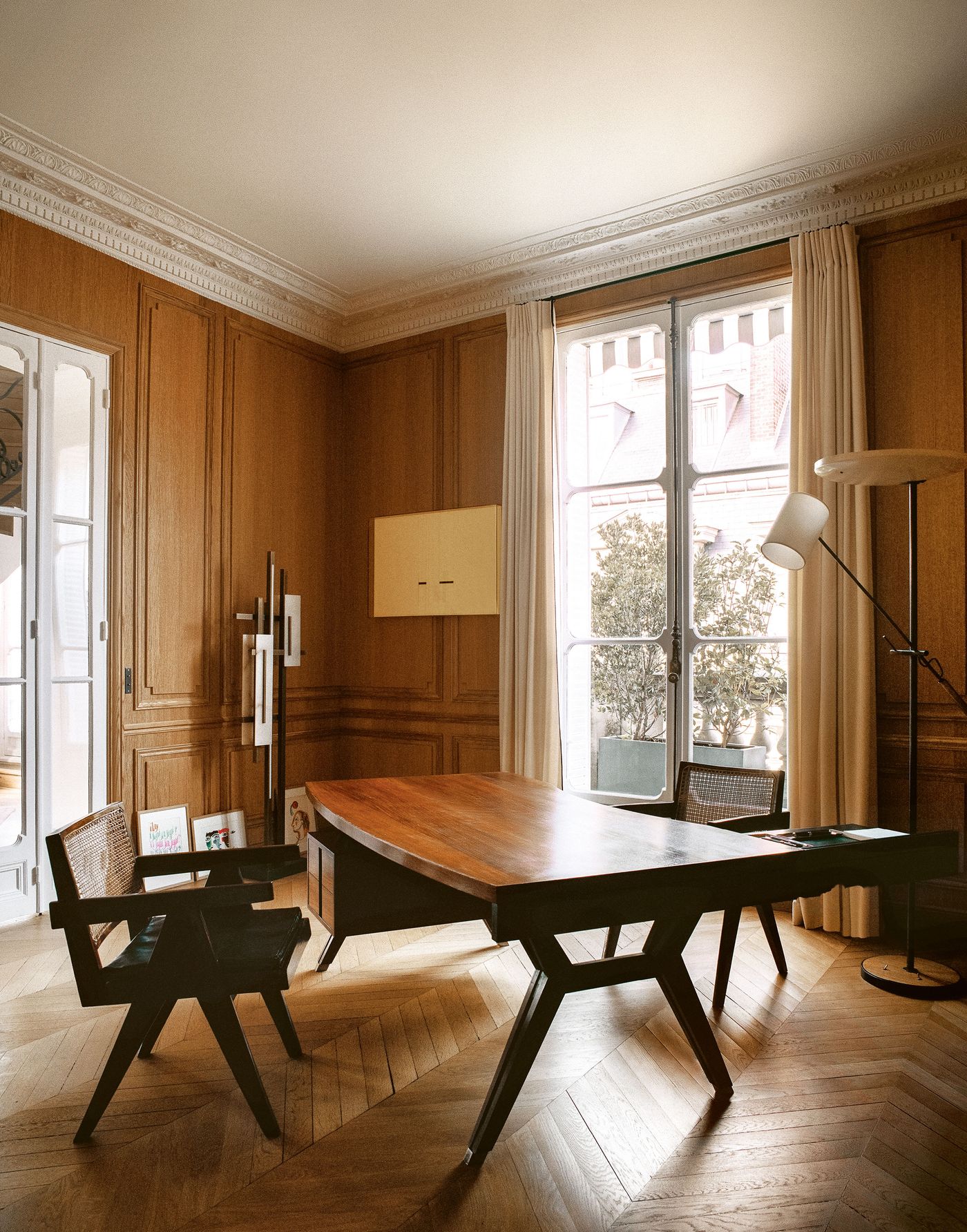 A stately home office with rich wooden paneling and a classic herringbone floor. A substantial dark wood desk with a unique base is central, accompanied by a cane-back chair, while expansive windows and a tall, slender modern lamp illuminate the elegant workspace.