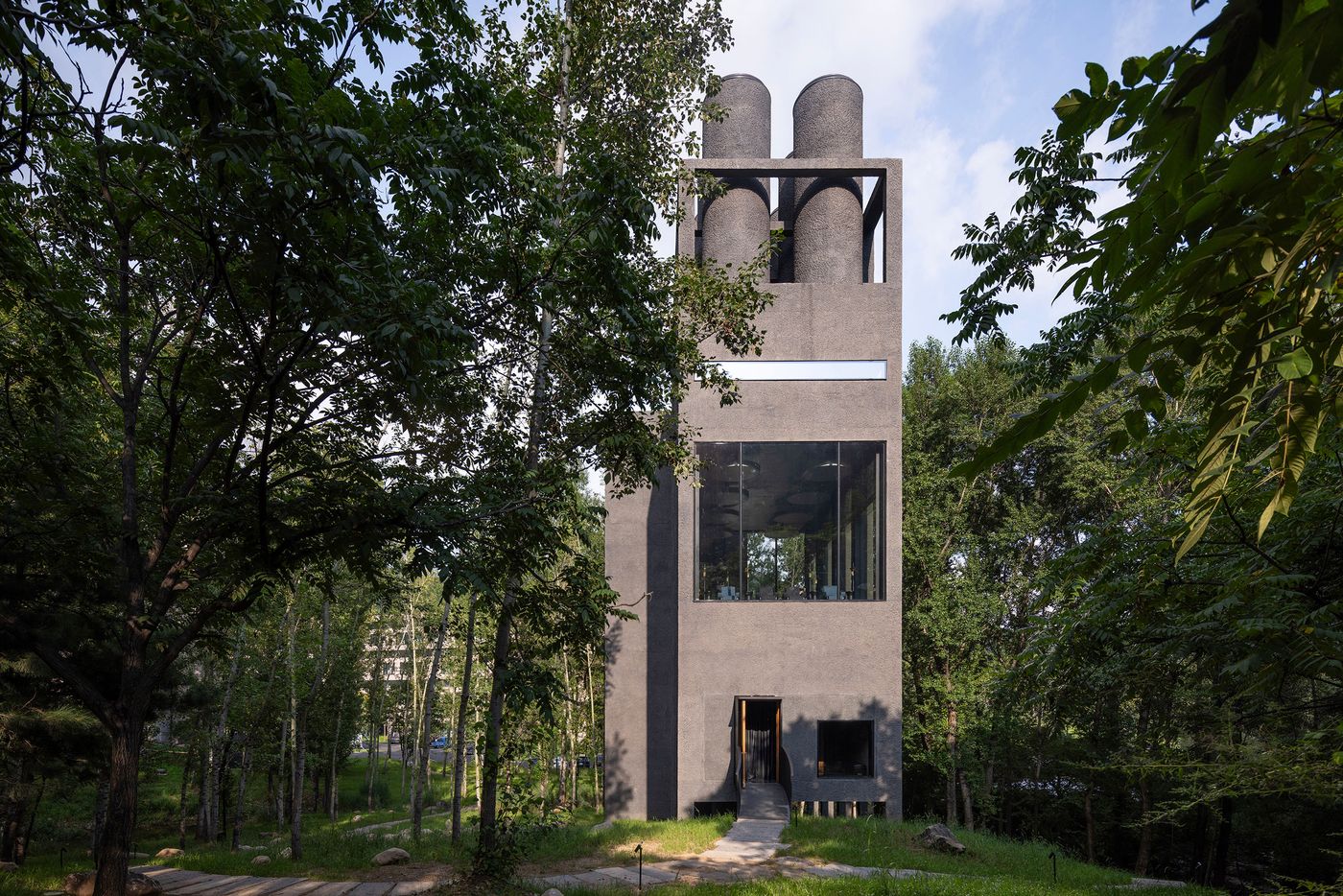 A modern, concrete building with three cylindrical structures on top, surrounded by lush green trees and a pathway leading to its entrance.
