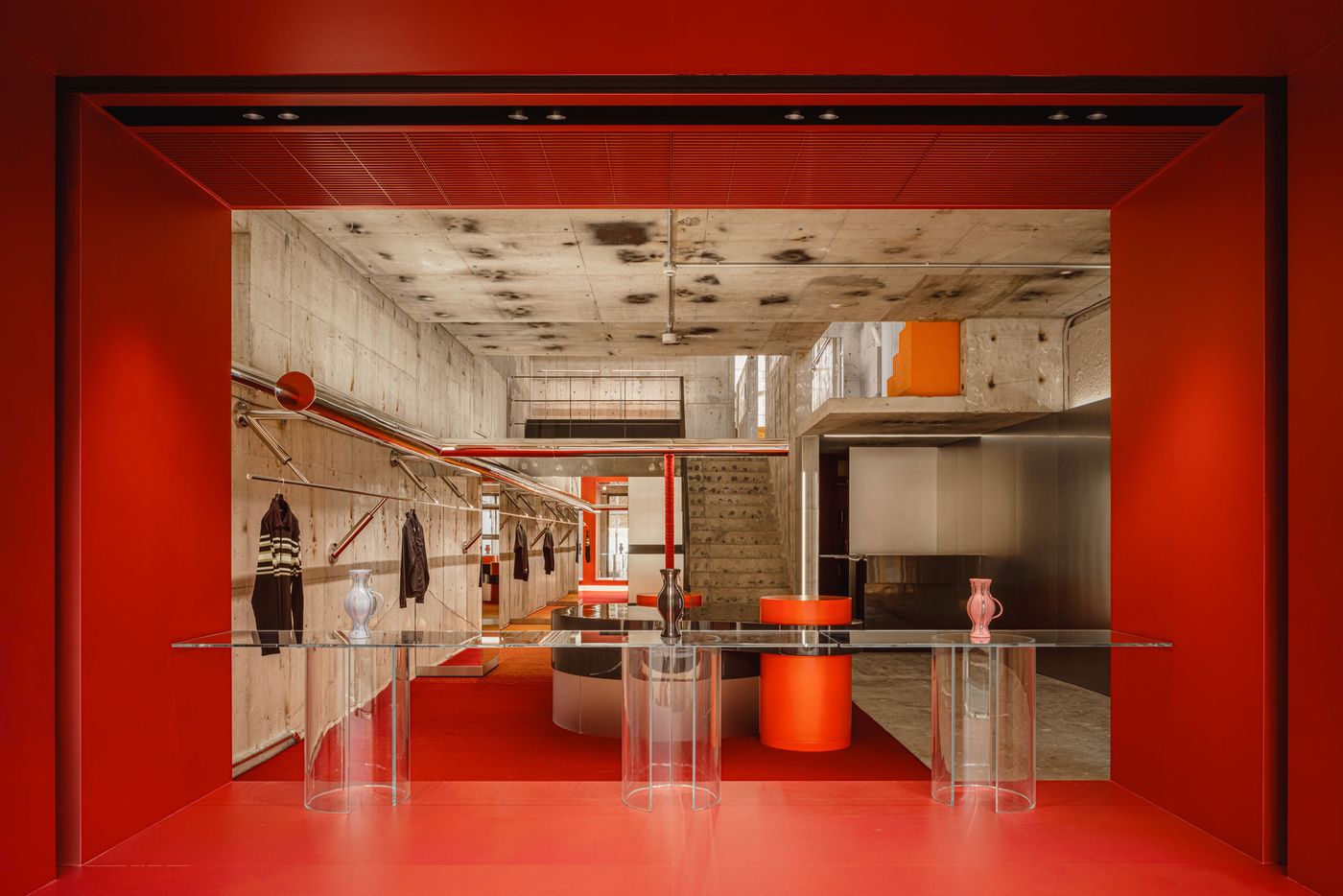 A vivid red portal frames the ground-floor interior like an oversized display window, focusing attention on the store’s central fixtures and long axial view. Transparent tables, cylindrical plinths and glossy black surfaces sit within the saturated enclosure, while rough concrete beyond heightens the contrast between raw architecture and polished retail design.