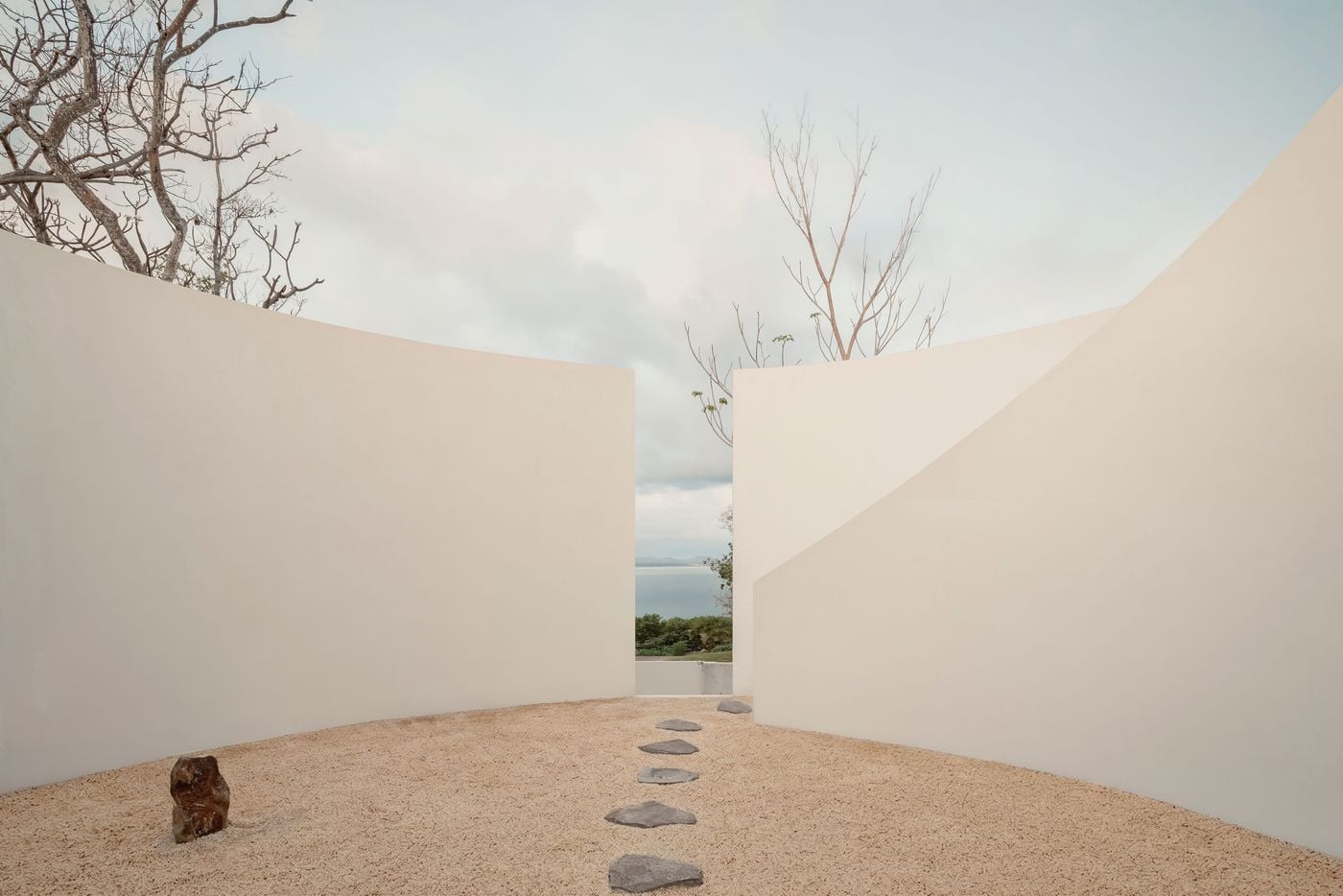 Curving cream walls part to frame a narrow vertical view of the bay and distant mountains, drawing the eye along a straight line of stepping stones across the gravel. A lone russet boulder and bare branches above the parapets lend the enclosure the stillness of a Japanese dry garden. Curving cream walls part to frame a narrow vertical view of the bay and distant mountains, drawing the eye along a straight line of stepping stones across the gravel. A lone russet boulder and bare branches above the parapets lend the enclosure the stillness of a Japanese dry garden.