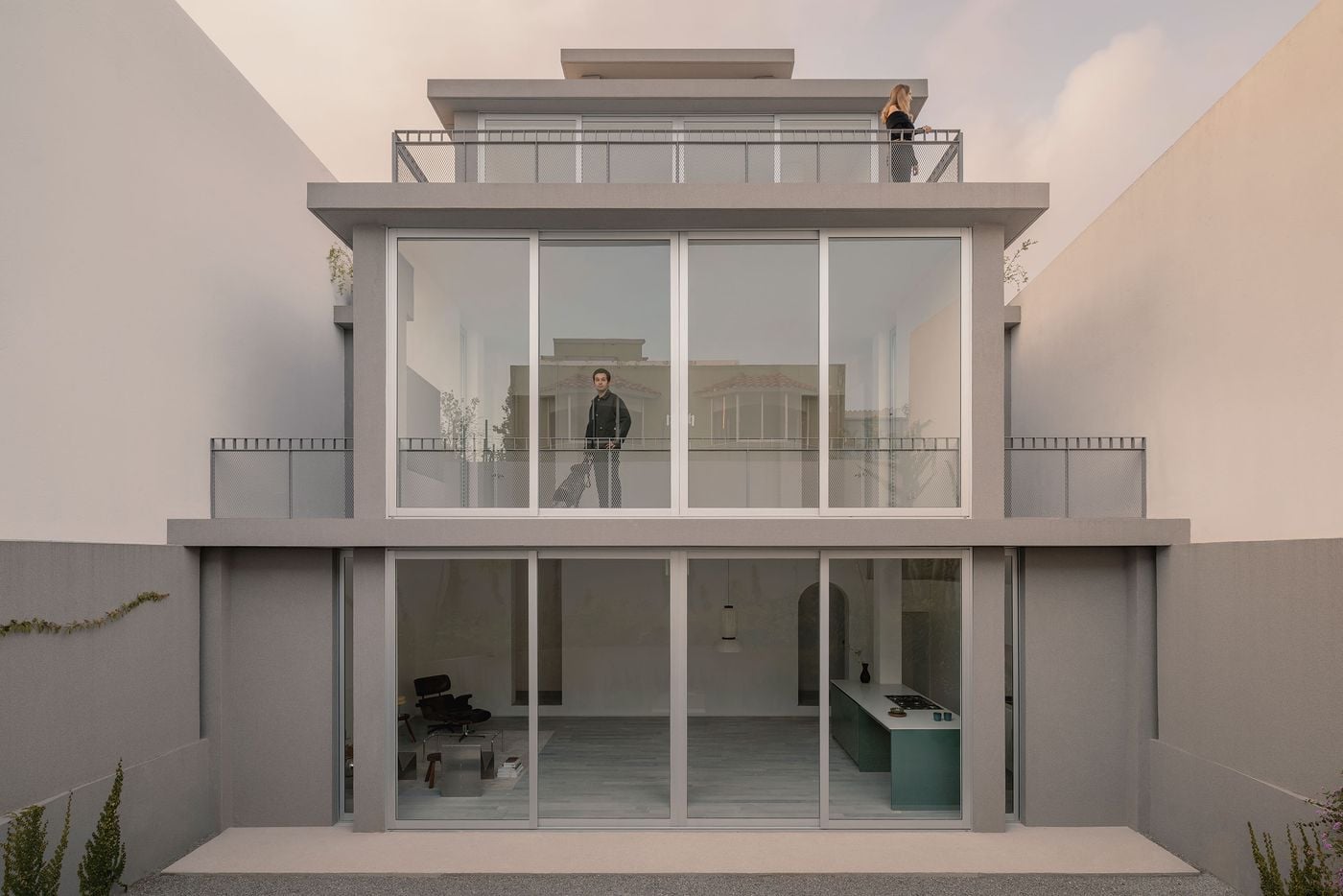 Casa Tlaloc’s rear elevation reveals the house as a sequence of progressively smaller glazed volumes rising above the courtyard. Wide sliding doors expose the minimalist ground-floor living space, while metal-mesh terraces wrap the upper levels, where two occupants animate the otherwise rigorous grey-and-glass composition. Casa Tlaloc’s rear elevation reveals the house as a sequence of progressively smaller glazed volumes rising above the courtyard. Wide sliding doors expose the minimalist ground-floor living space, while metal-mesh terraces wrap the upper levels, where two occupants animate the otherwise rigorous grey-and-glass composition.