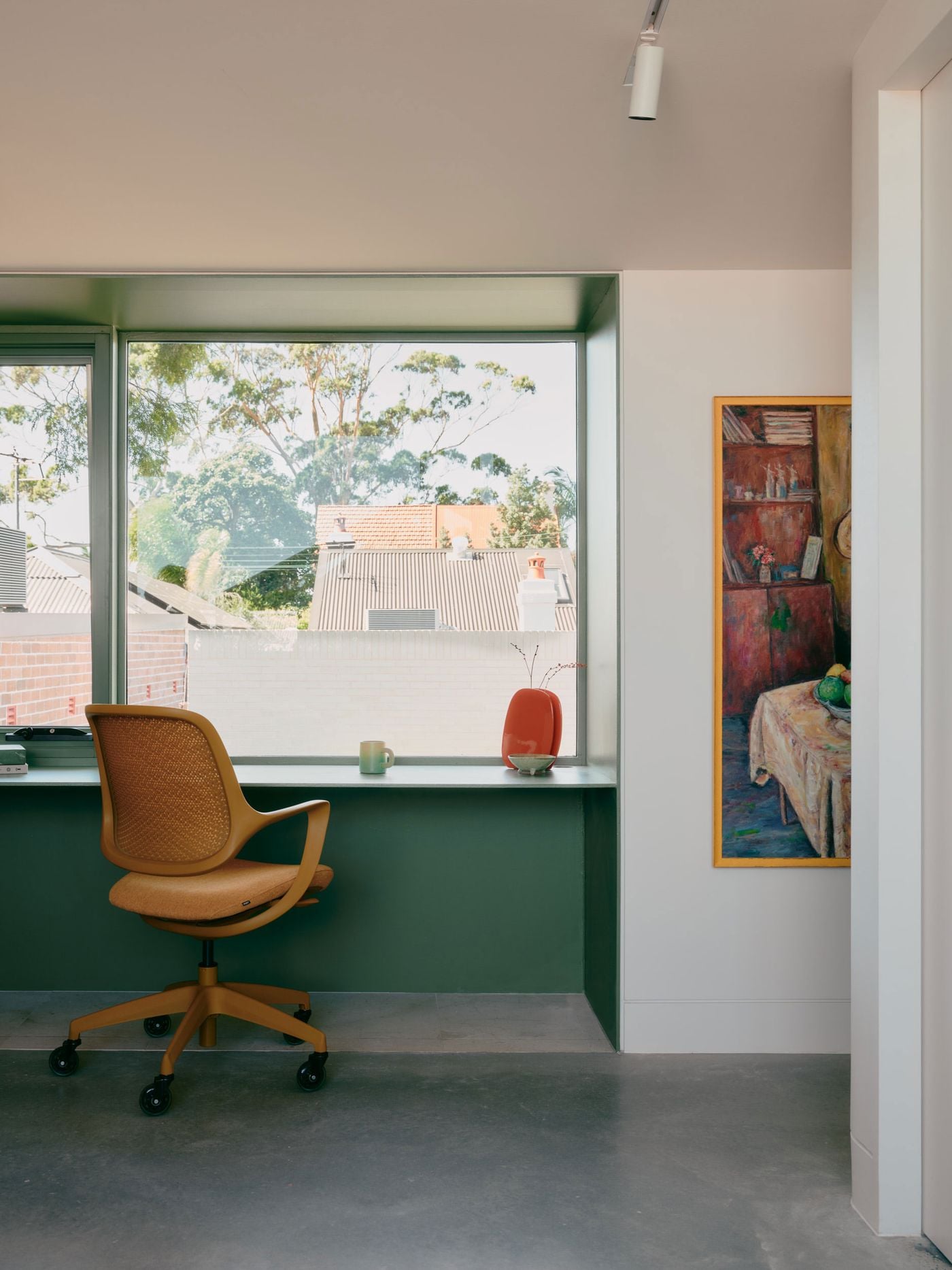 A built-in desk occupies the deep garden-facing window alcove, transforming the generous sill into a compact workspace. Sage-green joinery frames leafy suburban views, while a mustard-toned office chair and a warm-hued painting bring character to the otherwise pared-back interior of white walls and polished concrete flooring. A built-in desk occupies the deep garden-facing window alcove, transforming the generous sill into a compact workspace. Sage-green joinery frames leafy suburban views, while a mustard-toned office chair and a warm-hued painting bring character to the otherwise pared-back interior of white walls and polished concrete flooring.